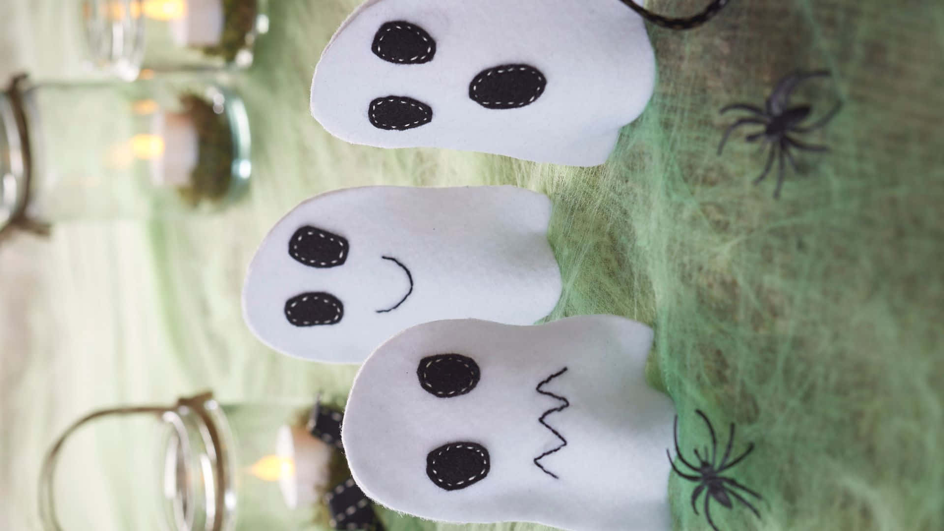Halloween Crafts 桌布 壁紙 — 三個手工製作的毛氈鬼魂，帶有縫製的臉孔，坐在綠色蜘蛛網布料上，旁邊有燃燒的蠟燭和黑色塑膠蜘蛛。