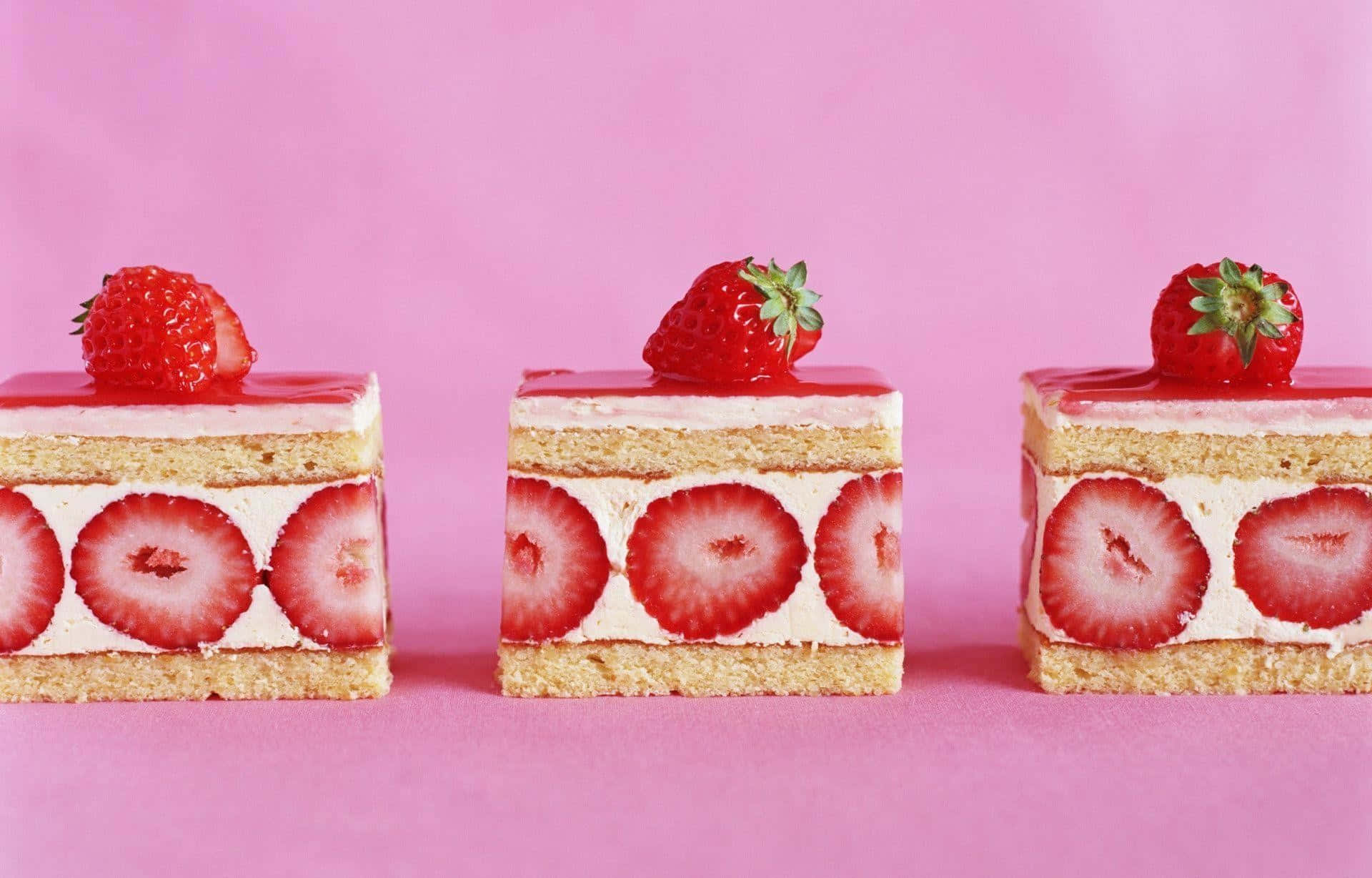 Kawaii Japanese Food 桌布 壁紙 — 三個方形草莓蛋糕，帶有光澤的粉紅色糖霜和新鮮的完整草莓在上面，置於純粉紅色背景上。