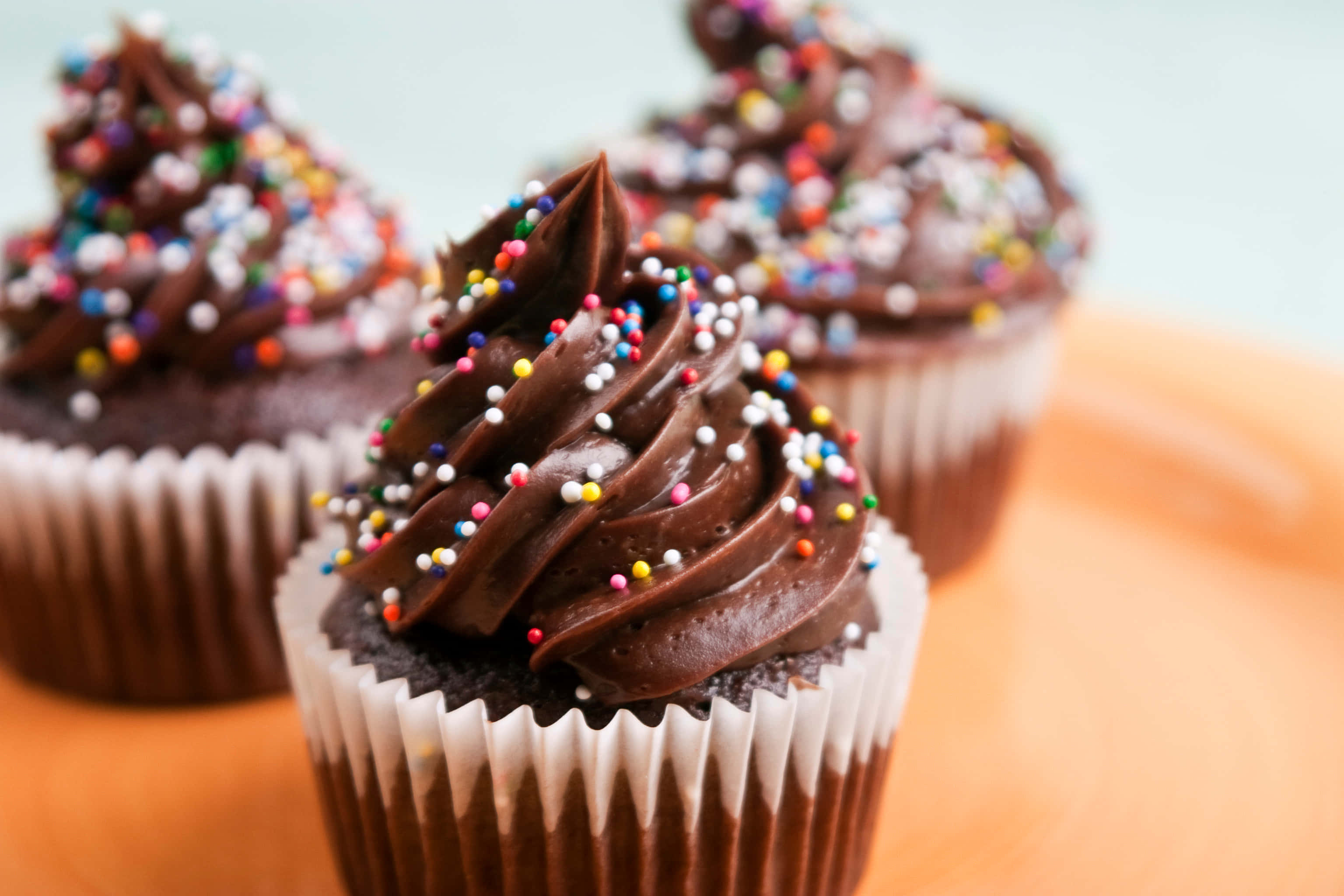 Cute Cupcake 桌布 壁紙 — 三個朱古力紙杯蛋糕，上面有濃郁的糖霜漩渦和色彩繽紛的糖粒，放在橙色碟上。