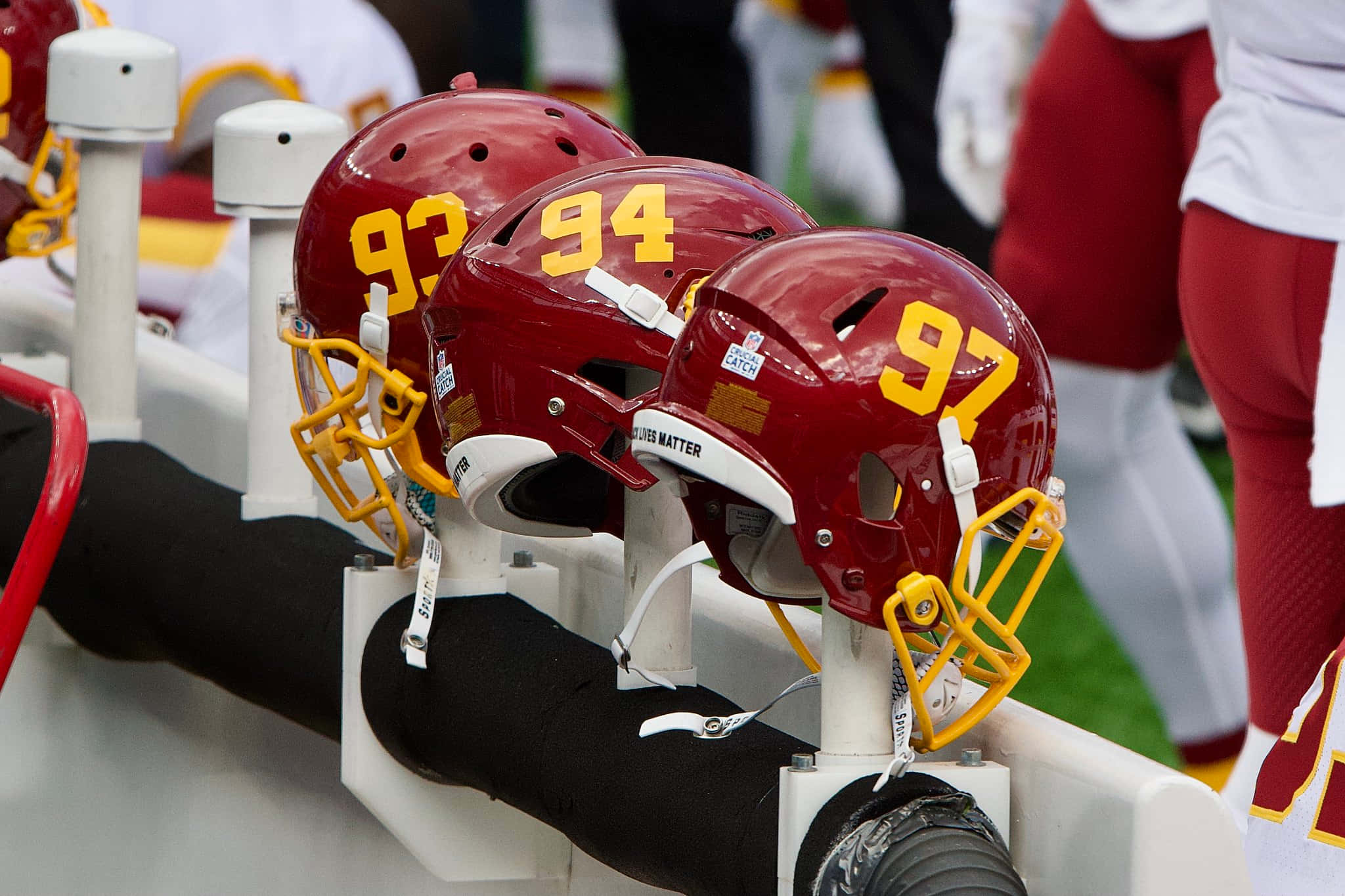Washington Football 桌布 壁紙 — 三個栗色美式足球頭盔，帶有黃色數字93、94和97，放在白色支架上，旁邊是穿著紅色球衣的球員。