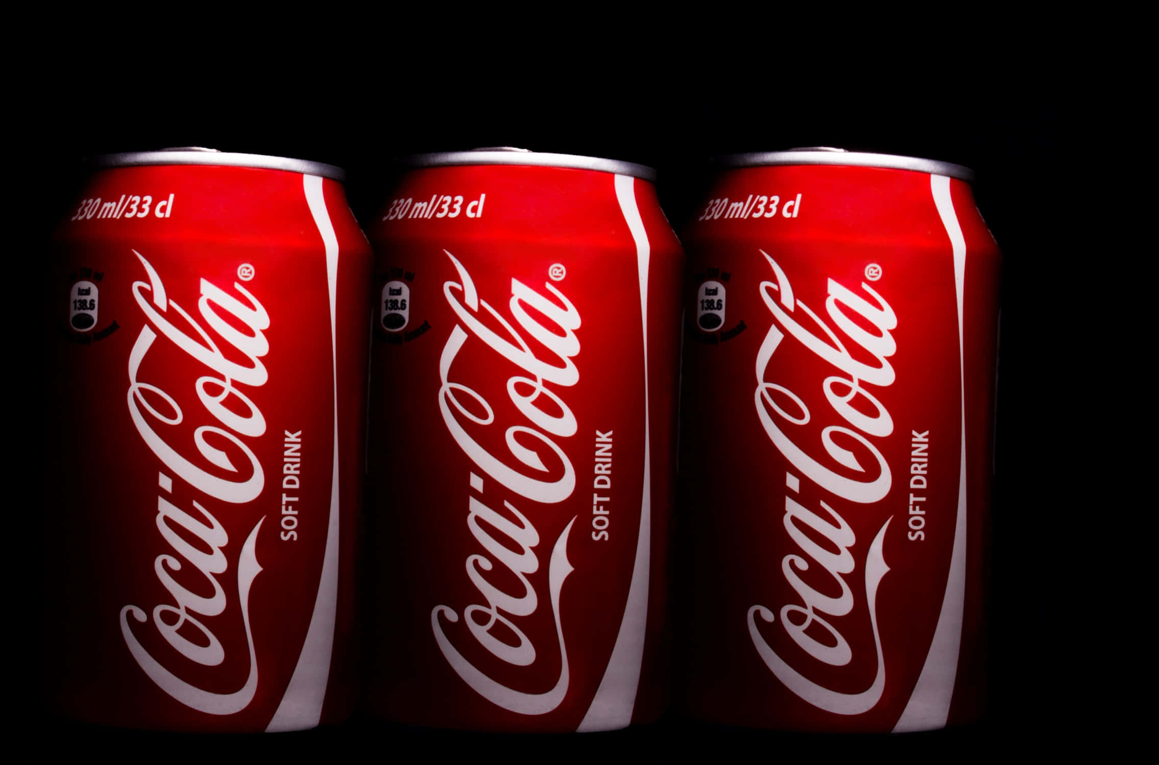 coca cola 桌布 壁紙 — 三個標誌性的紅色可口可樂罐，帶有白色字體，排成一排，背景為純黑色。