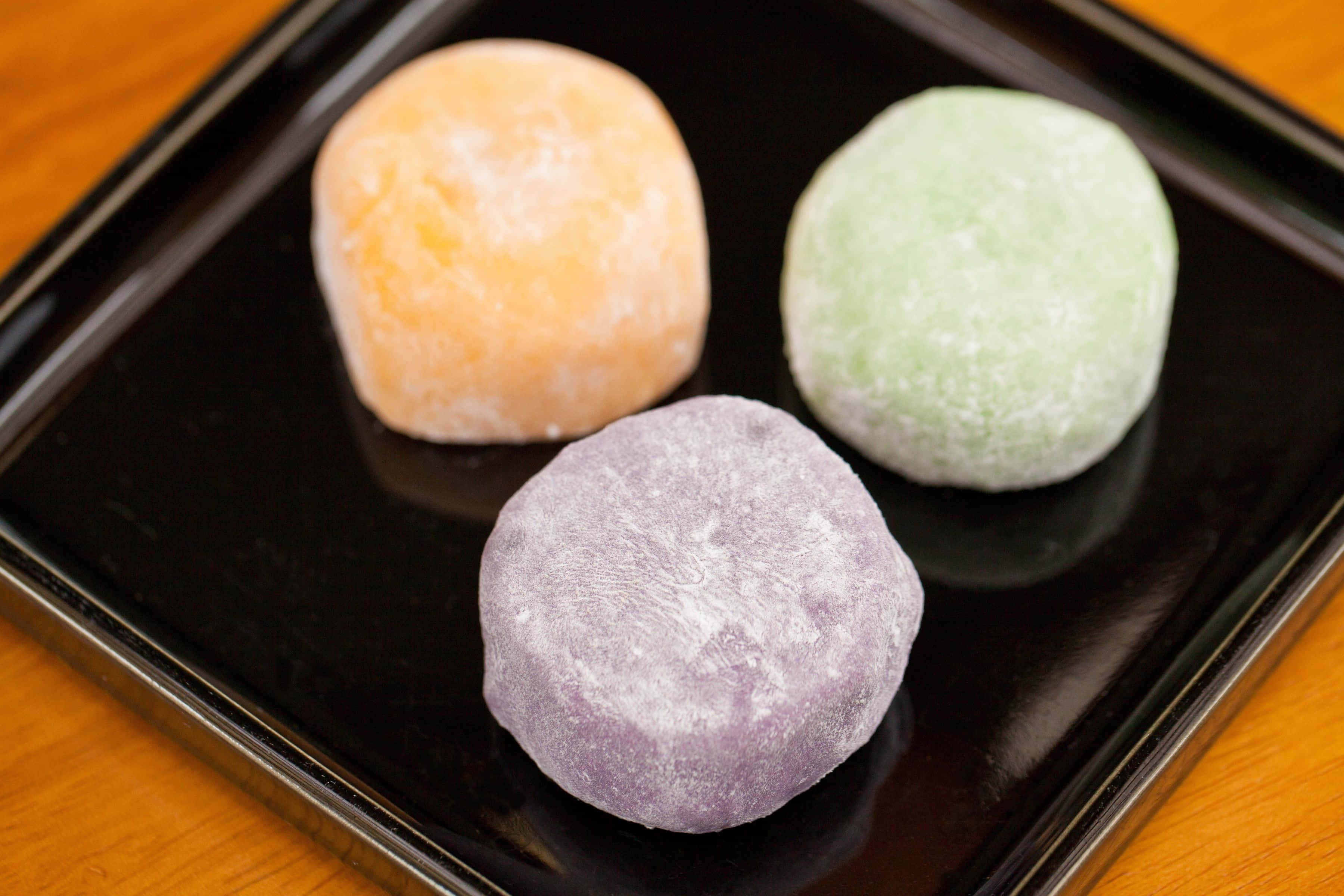 Japanese Sweets 桌布 壁紙 — 三個沾滿粉末的麻糬糰子，分別為橙色、綠色和紫色，放在溫暖木質表面上的亮面黑色托盤上。