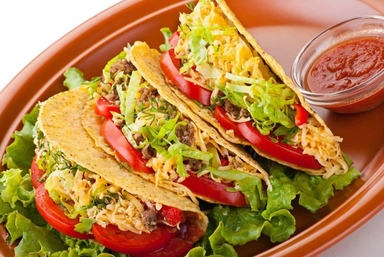 Taco 桌布 壁紙 — 三個滿載牛肉的玉米餅，搭配生菜、番茄和芝士，放在蔬菜床上，旁邊有一小碗莎莎醬。