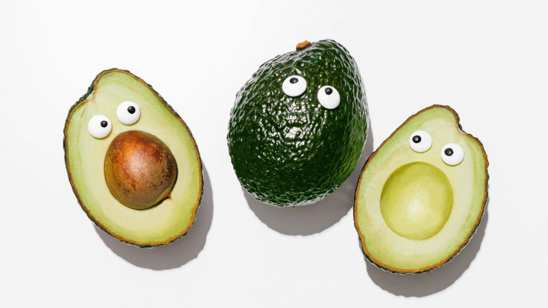 Cute Avocado 桌布 壁紙 — 三個牛油果帶有咕嚕眼睛，排列在純白色背景上，一個完整，一個切半有核，一個切半空心。