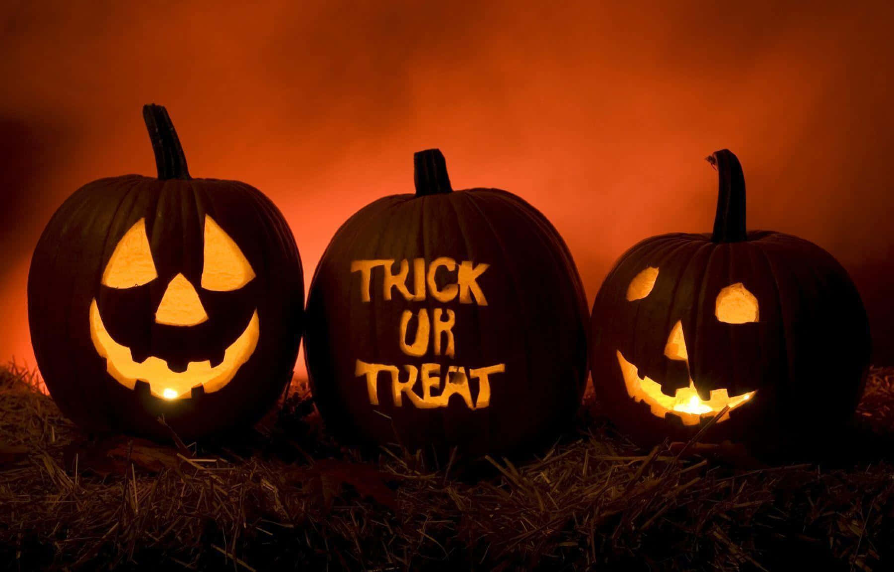 Halloween Props 桌布 壁紙 — 三個發光的傑克南瓜燈，帶有雕刻的臉孔和「不給糖就搗蛋」文字，坐在乾草上，背景是煙霧般的橙色。