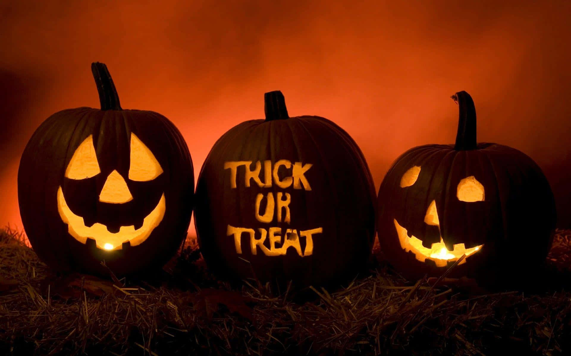 Trick-or-treaters 桌布 壁紙 — 三個發光的傑克南瓜燈，帶有雕刻的臉孔和「trick or treat」文字，放在乾草上，背景是煙霧瀰漫的橙色。