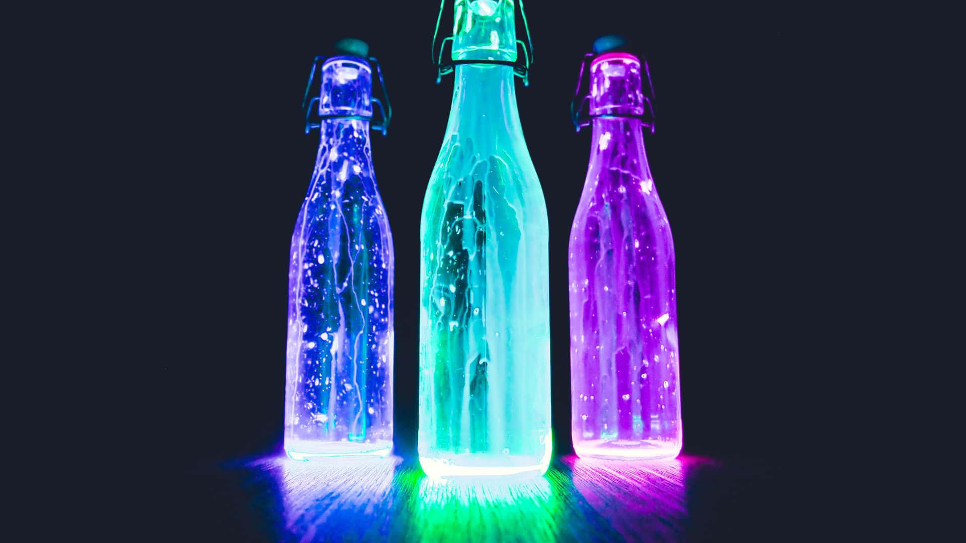 Glow In The Dark 桌布 壁紙 — 三個發光的玻璃瓶裝滿閃爍的液體，在黑暗背景下散發出鮮豔的藍色、綠色和紫色光芒。