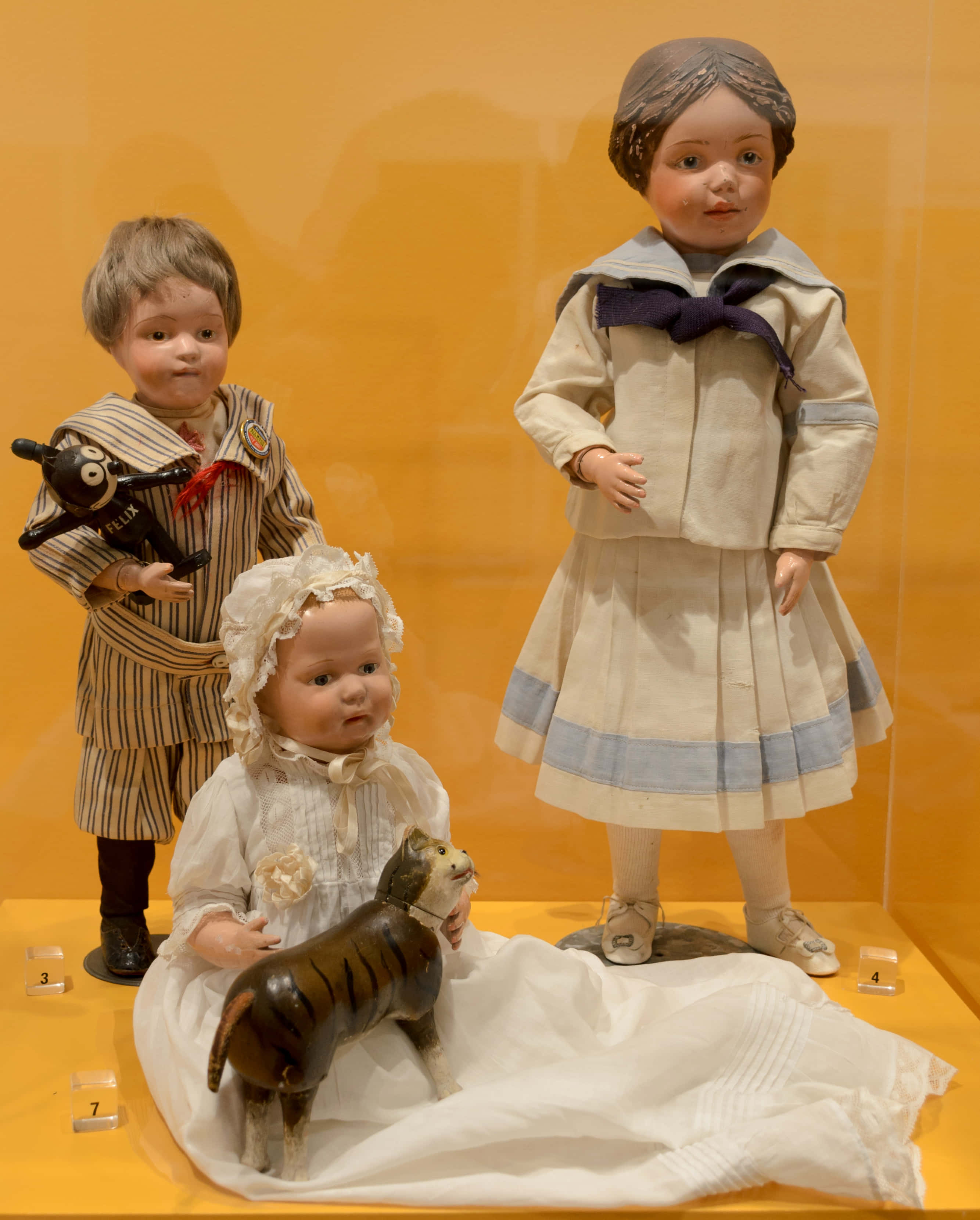 Dolls 桌布 壁紙 — 三個穿著時代服裝的復古娃娃站在博物館展示櫃中，背景是溫暖的黃色牆壁。