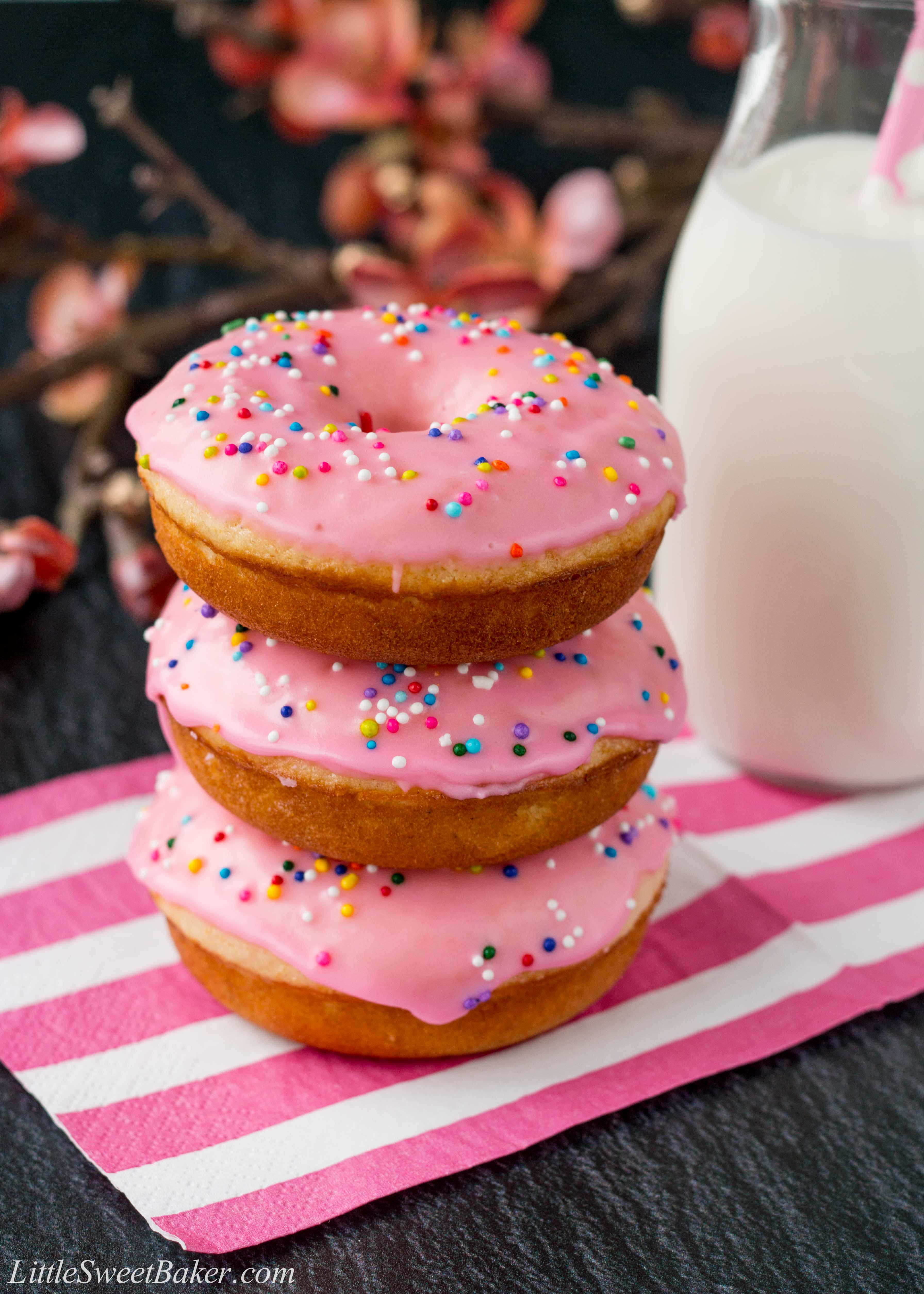 Glazed Donut 桌布 壁紙 — 三個粉紅色糖霜甜甜圈，上面有彩色糖粒，疊放在粉白條紋餐巾上，旁邊有一杯牛奶。