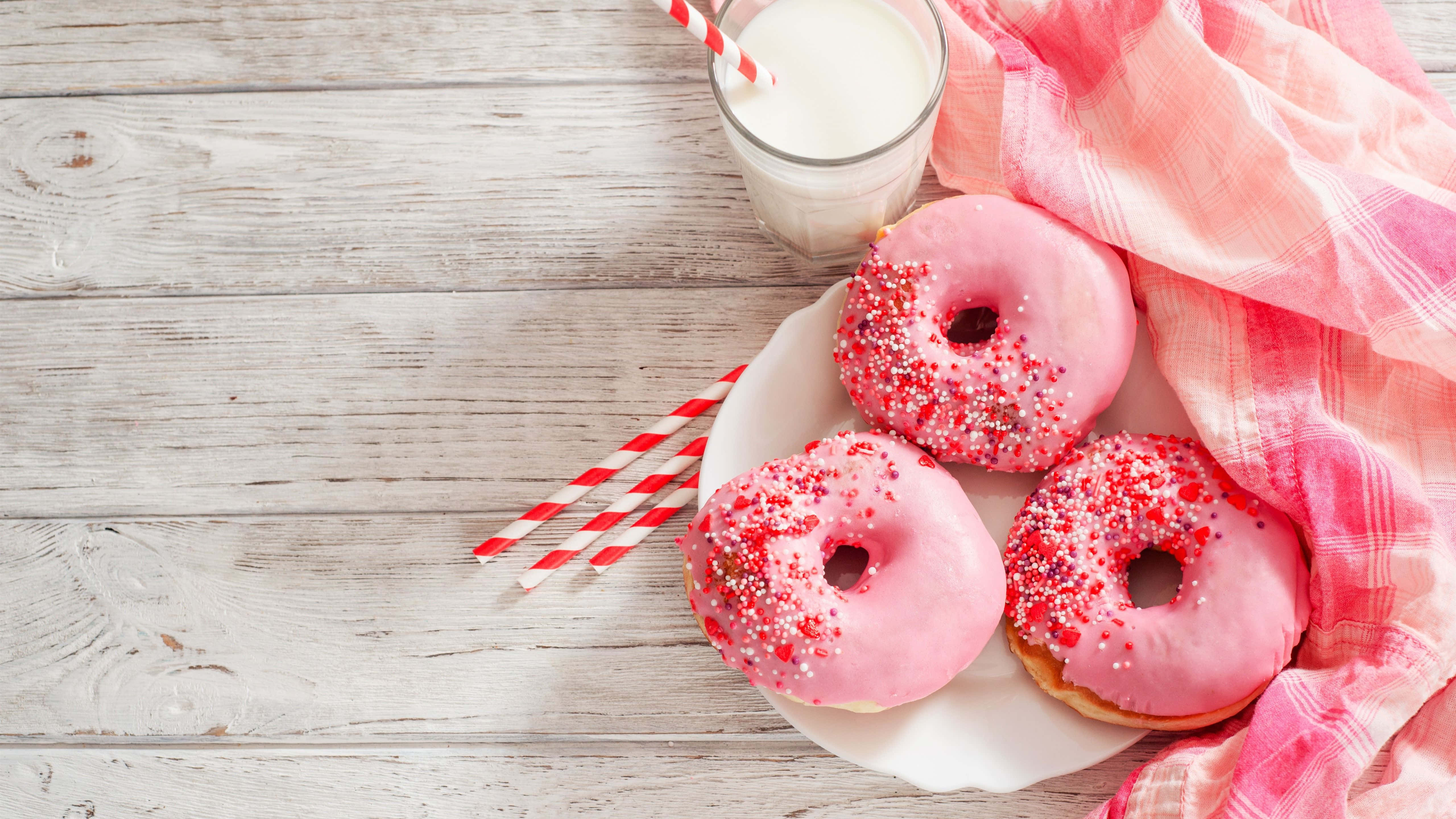 Cute Donut 桌布 壁紙 — 三個粉紅色糖霜甜甜圈配紅色糖粒，放在白色碟上，旁邊有一杯牛奶和一塊粉紅色布，置於風化的木桌上。