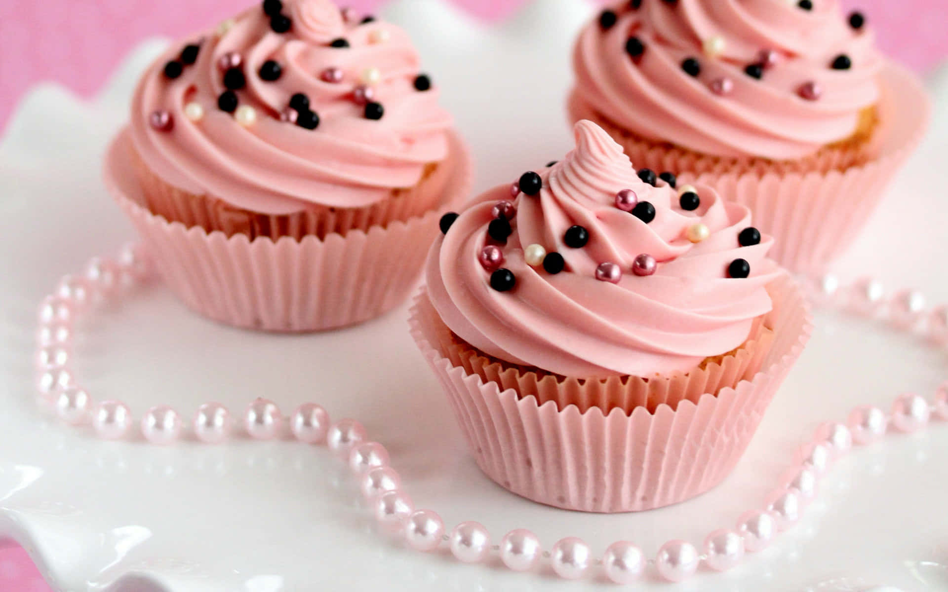 Cute Cupcake 桌布 壁紙 — 三個粉紅色糖霜紙杯蛋糕，上面有黑色和珍珠色糖粒，放在白色碟子上，珍珠頸鏈形成心形。