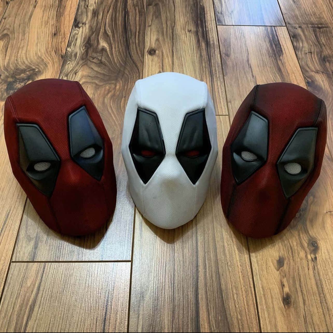 Deadpool Mask 桌布 壁紙 — 三個紅色和白色的死侍面具並排放在有明顯木紋的木地板上。
