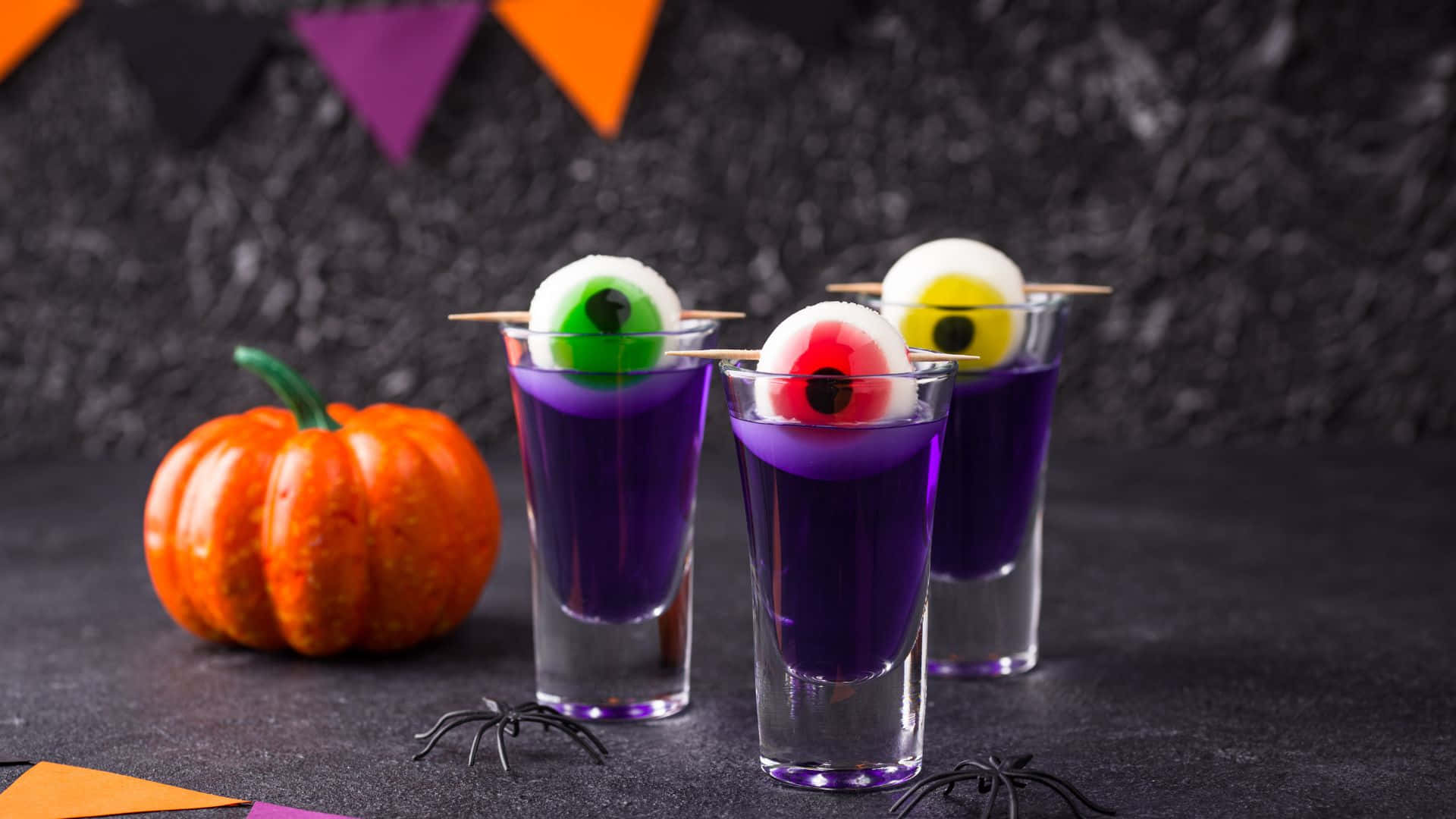 Spooky Cocktails 桌布 壁紙 — 三個紫色萬聖節小酒杯，上面放著糖果眼球，旁邊有一個小南瓜和黑色蜘蛛裝飾。