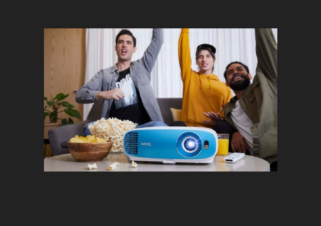 Home Entertainment 桌布 壁紙 — 三個興奮的朋友在沙發上歡呼，藍色投影機將光線投射到牆上，附近有爆谷和飲料。