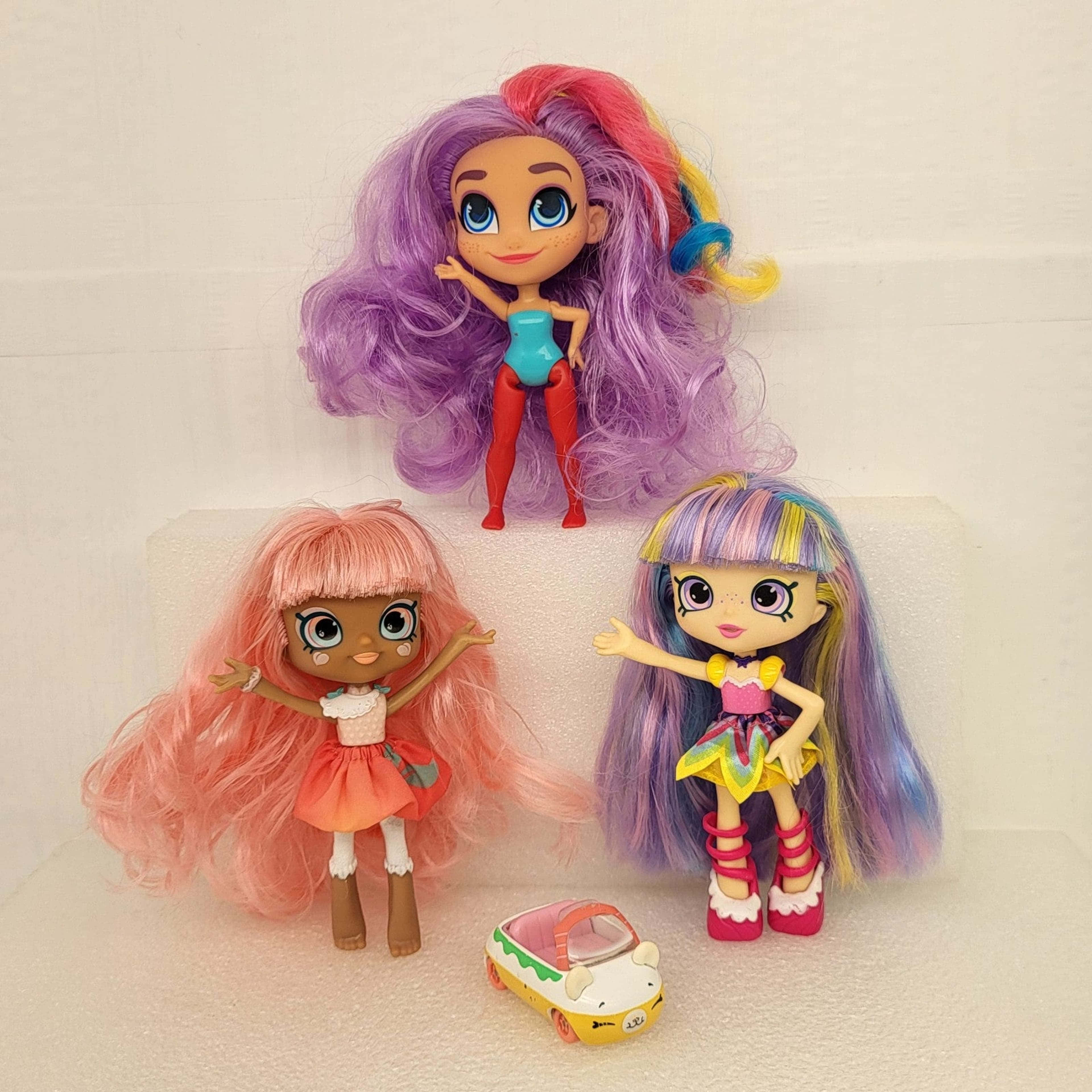 Shopkins 桌布 壁紙 — 三個色彩繽紛、擁有鮮豔彩虹頭髮的洋娃娃，在白色紋理表面上，開心地在一輛小型玩具車旁邊擺姿勢。