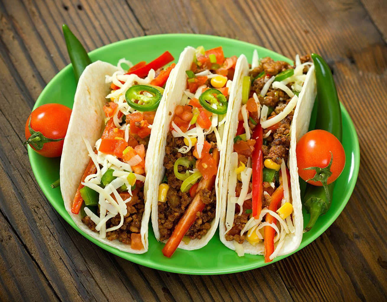 Taco 桌布 壁紙 — 三個色彩繽紛的墨西哥卷餅，餡料有肉、芝士和蔬菜，放在一個亮綠色的碟子上，置於鄉村風格的木桌上。