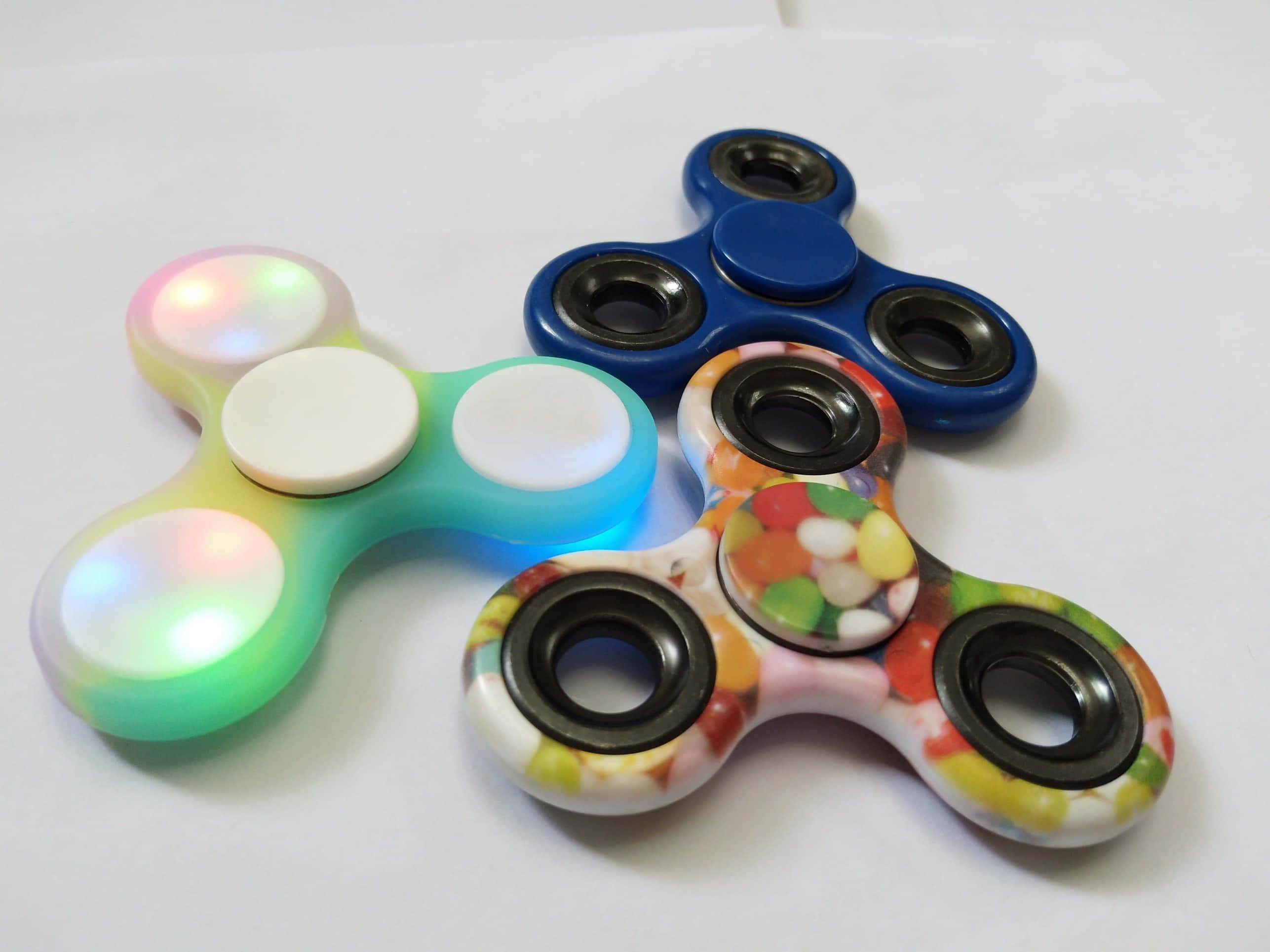Fidget 桌布 壁紙 — 三個色彩繽紛的指尖陀螺放在白色表面上，一個發出彩虹光芒，一個藍色，一個糖果圖案。