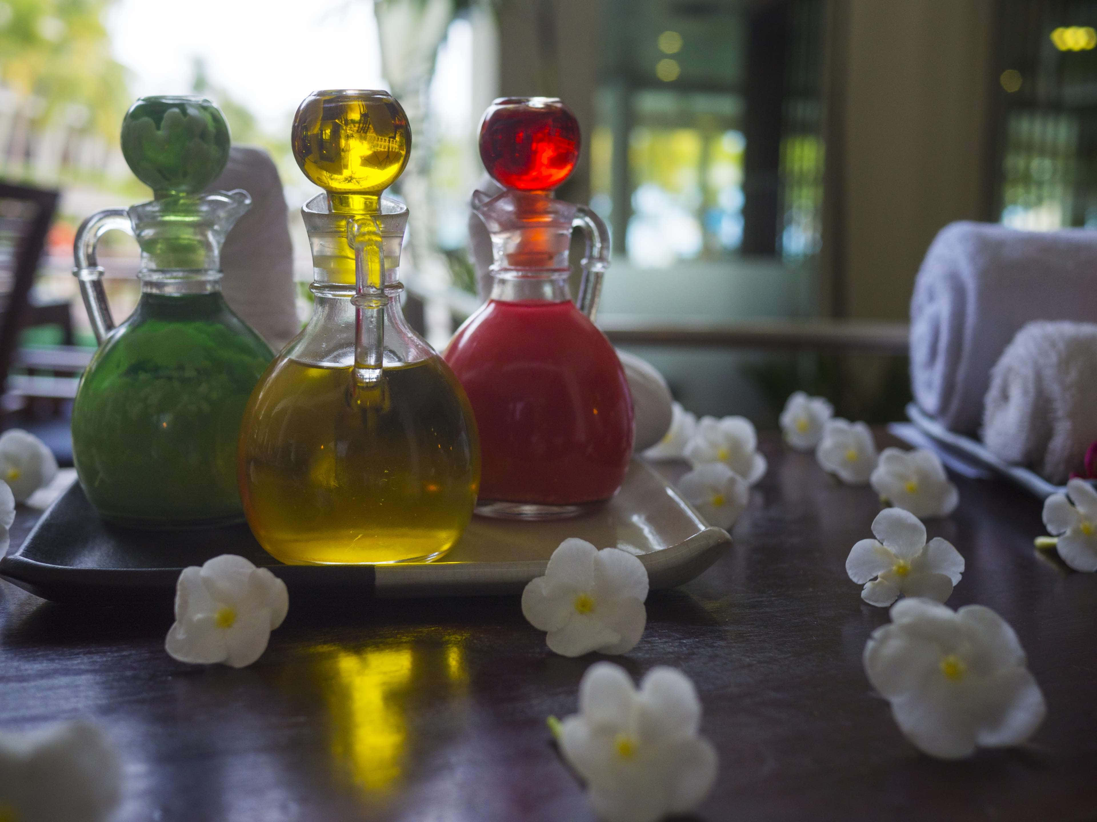 Aromatherapy 桌布 壁紙 — 三個色彩繽紛的玻璃瓶，裝有綠色、黃色和紅色的液體，放在一個托盤上，周圍環繞著白色花朵，置於深色木桌上。