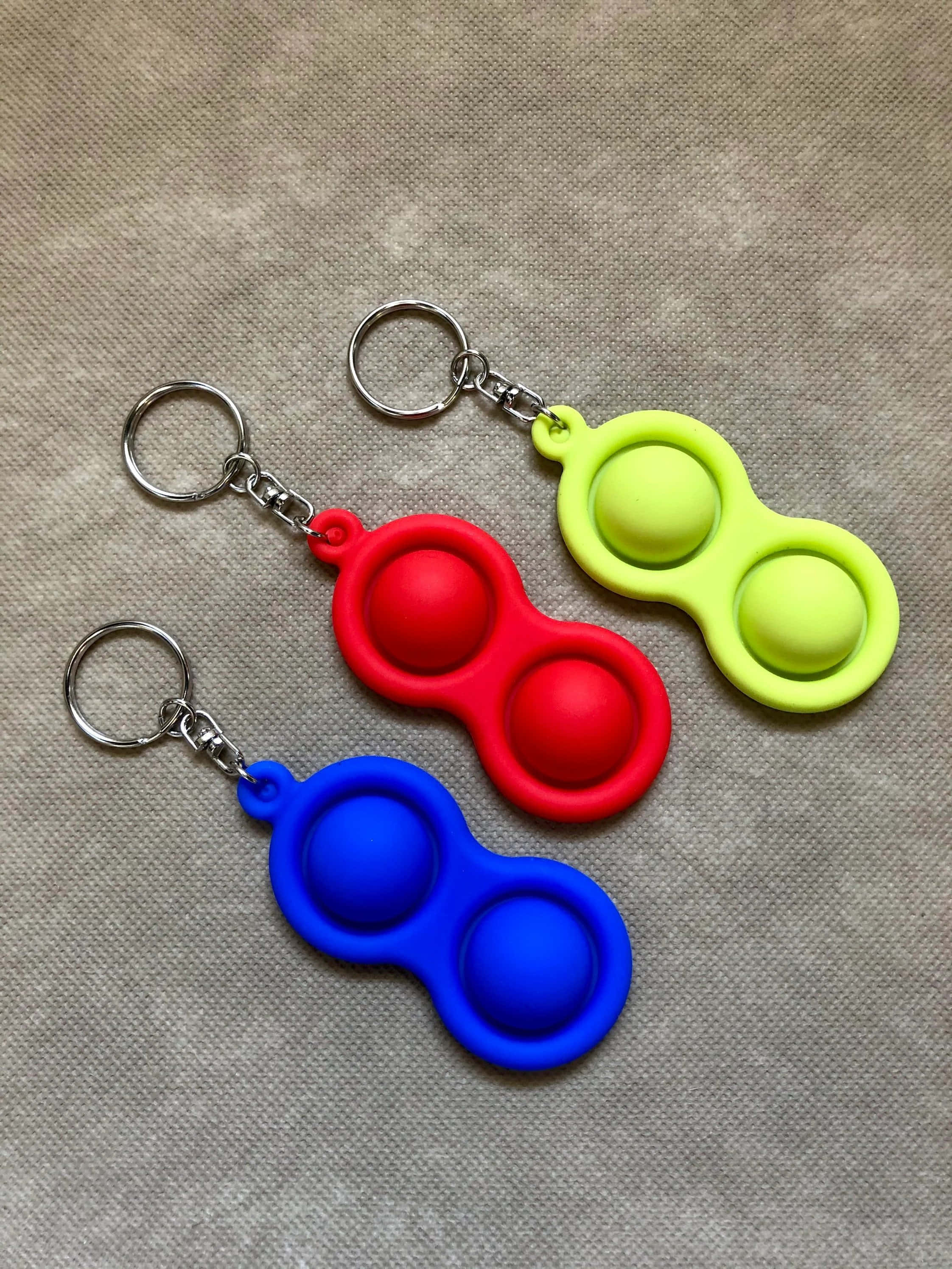 Fidget 桌布 壁紙 — 三個色彩繽紛的矽膠彈力減壓鎖匙扣，分別為紅色、藍色和青檸綠色，放置在帶有紋理的米色布料表面上。