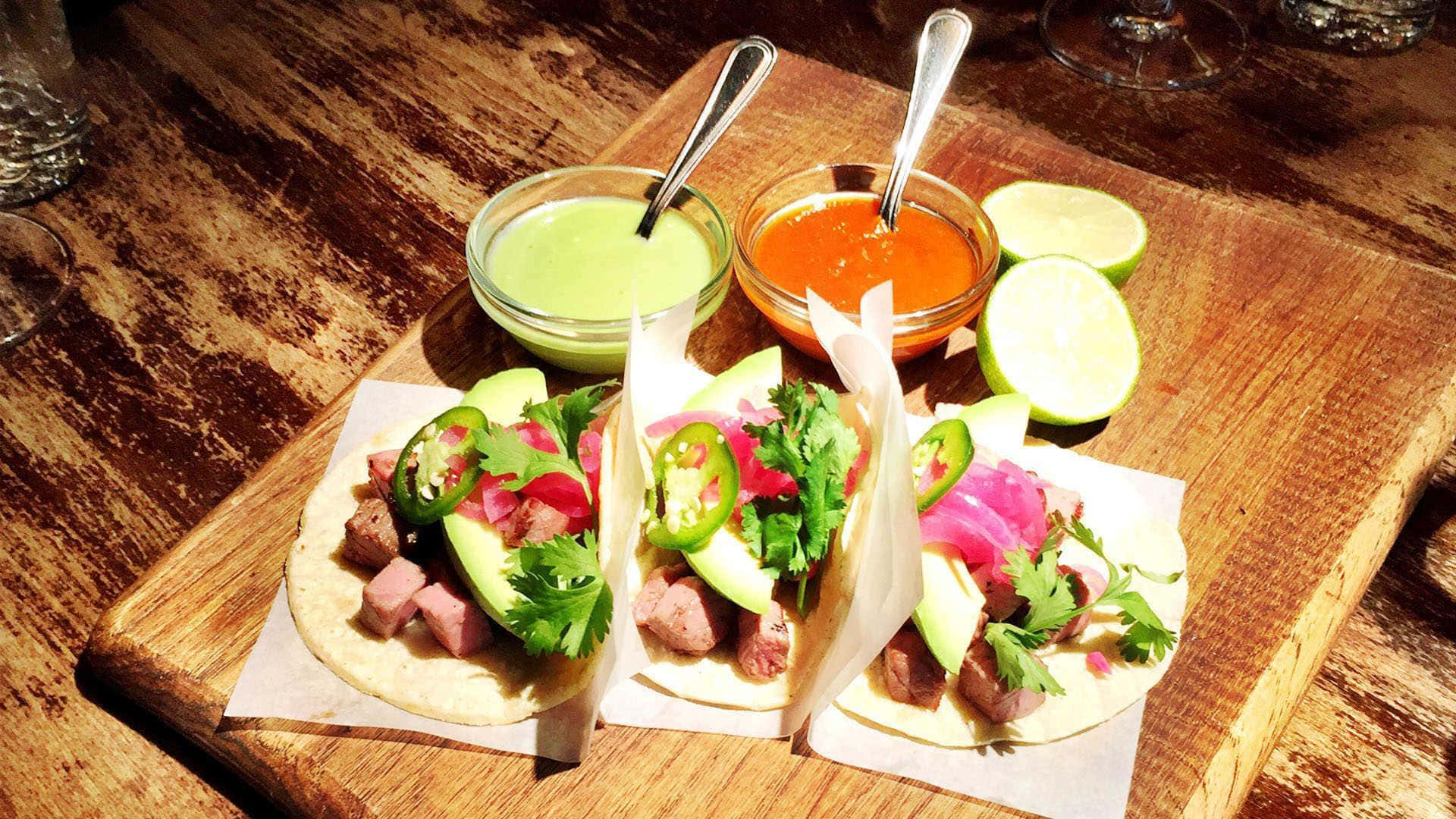 Taco 桌布 壁紙 — 三個色彩鮮豔的墨西哥卷餅，搭配牛油果、醃洋蔥和墨西哥辣椒，放在質樸的木板上，旁邊有青檸角和色彩繽紛的莎莎醬。