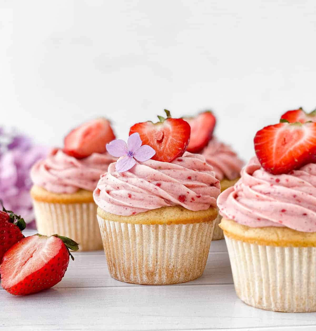 Pink Cupcakes 桌布 壁紙 — 三個草莓杯子蛋糕，配上粉紅色糖霜，上面放上新鮮切片草莓和一朵精緻的紫色花，放在白色木質表面上。