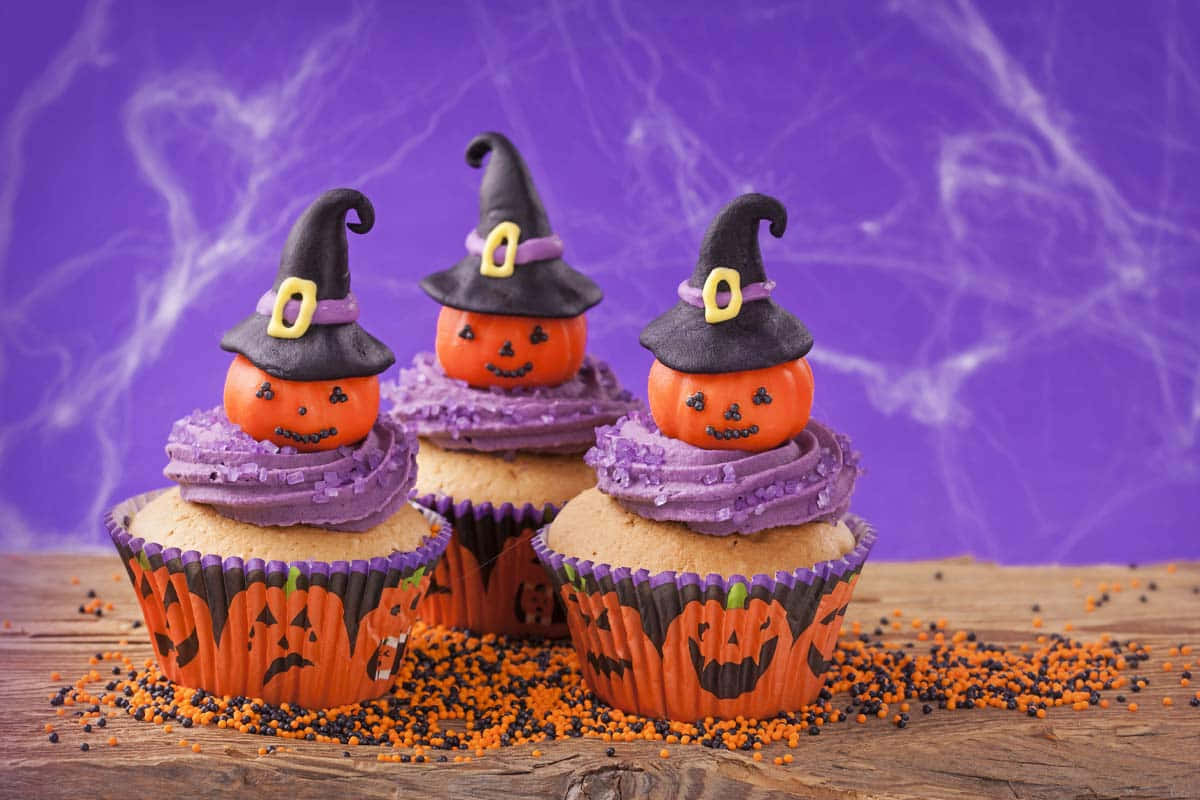 Halloween Cupcakes 桌布 壁紙 — 三個萬聖節紙杯蛋糕，頂部有戴著巫婆帽的傑克南瓜燈，放在木質表面上，撒有橙色和黑色糖粒，背景是紫色蜘蛛網。