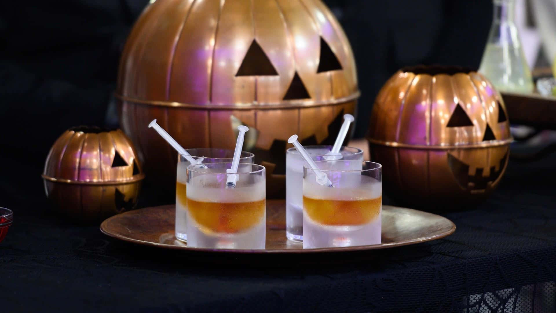 Spooky Cocktails 桌布 壁紙 — 三個萬聖節雞尾酒杯放在銅托盤上，旁邊是發出紫色光芒的金屬南瓜燈。