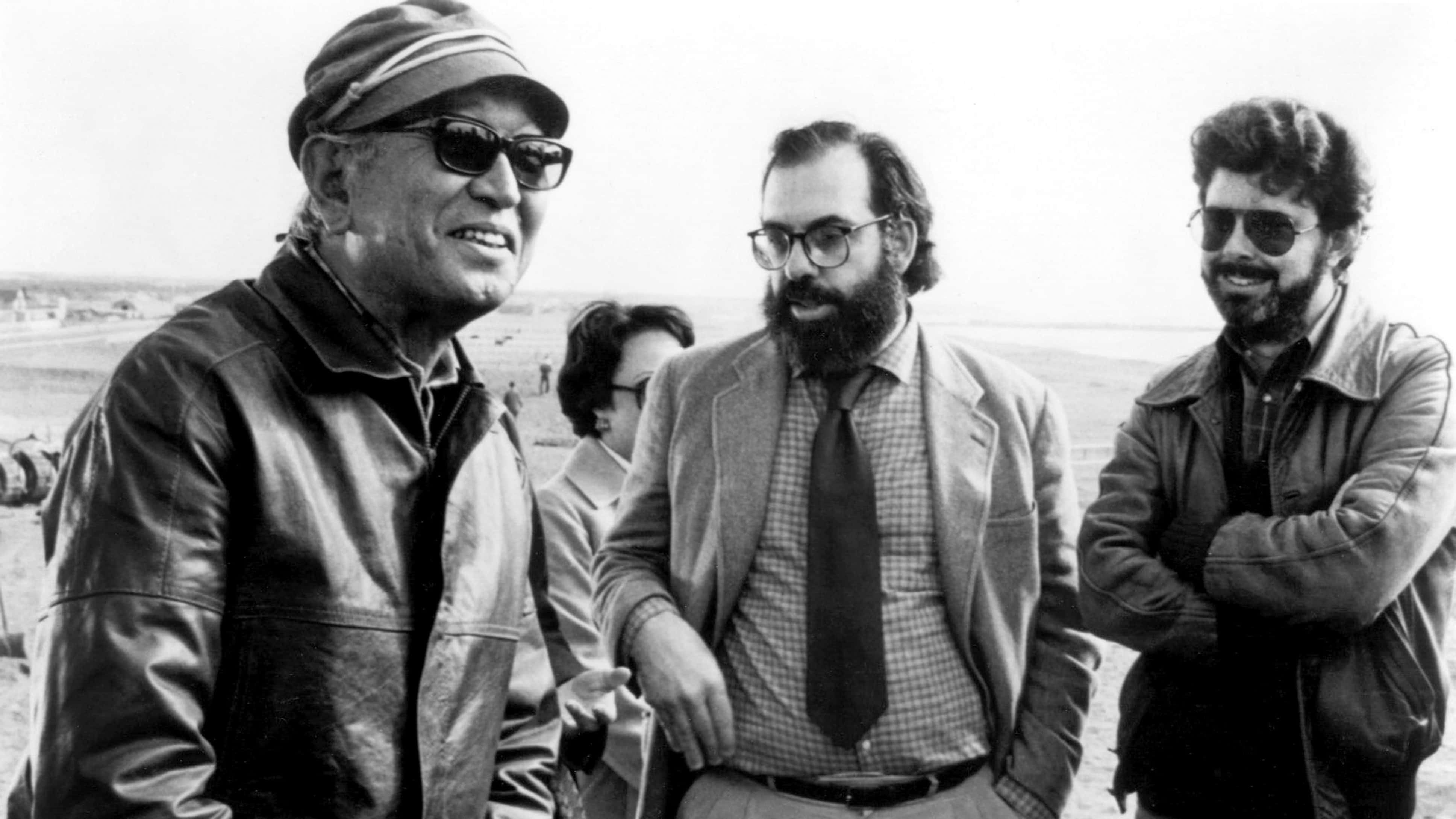 Akira Kurosawa 桌布 壁紙 — 三個身穿外套和太陽眼鏡的男人一起站在明亮的沙灘上，背後是遙遠的海岸線。