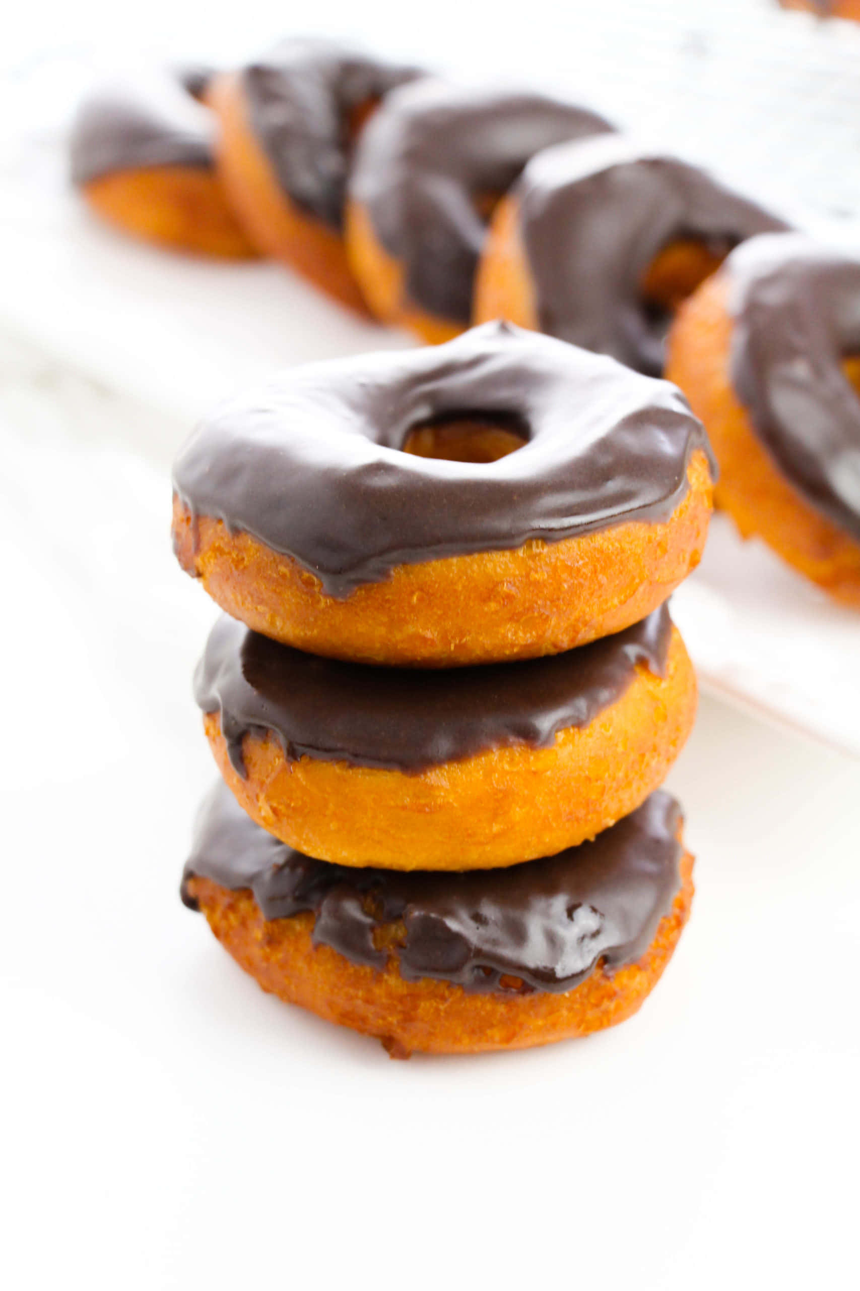 Glazed Donut 桌布 壁紙 — 三個金棕色朱古力釉面冬甩堆疊在焦點中，背景有更多模糊的冬甩在白色表面上。