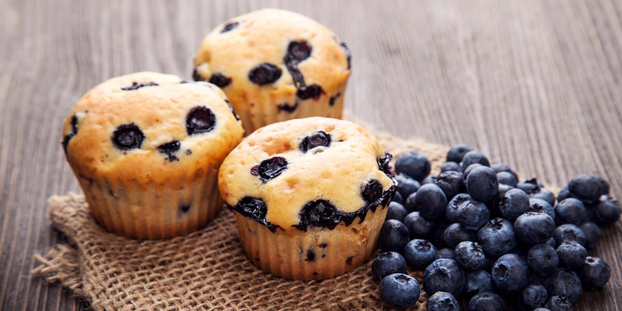 Blueberry Muffins 桌布 壁紙 — 三個金黃色的藍莓鬆餅配新鮮藍莓，放在木頭上的粗麻布上，旁邊是一堆飽滿的藍莓。