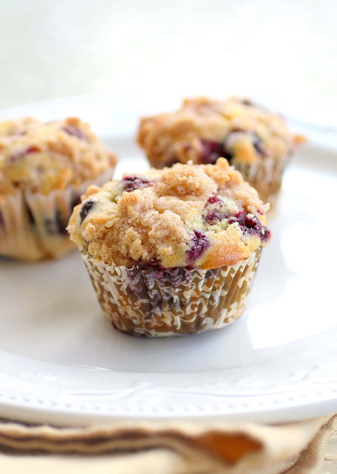 Blueberry Muffins 桌布 壁紙 — 三個金黃色藍莓鬆餅，上面有酥脆的奶酥粒，放在白色碟子上，配以米色餐巾。