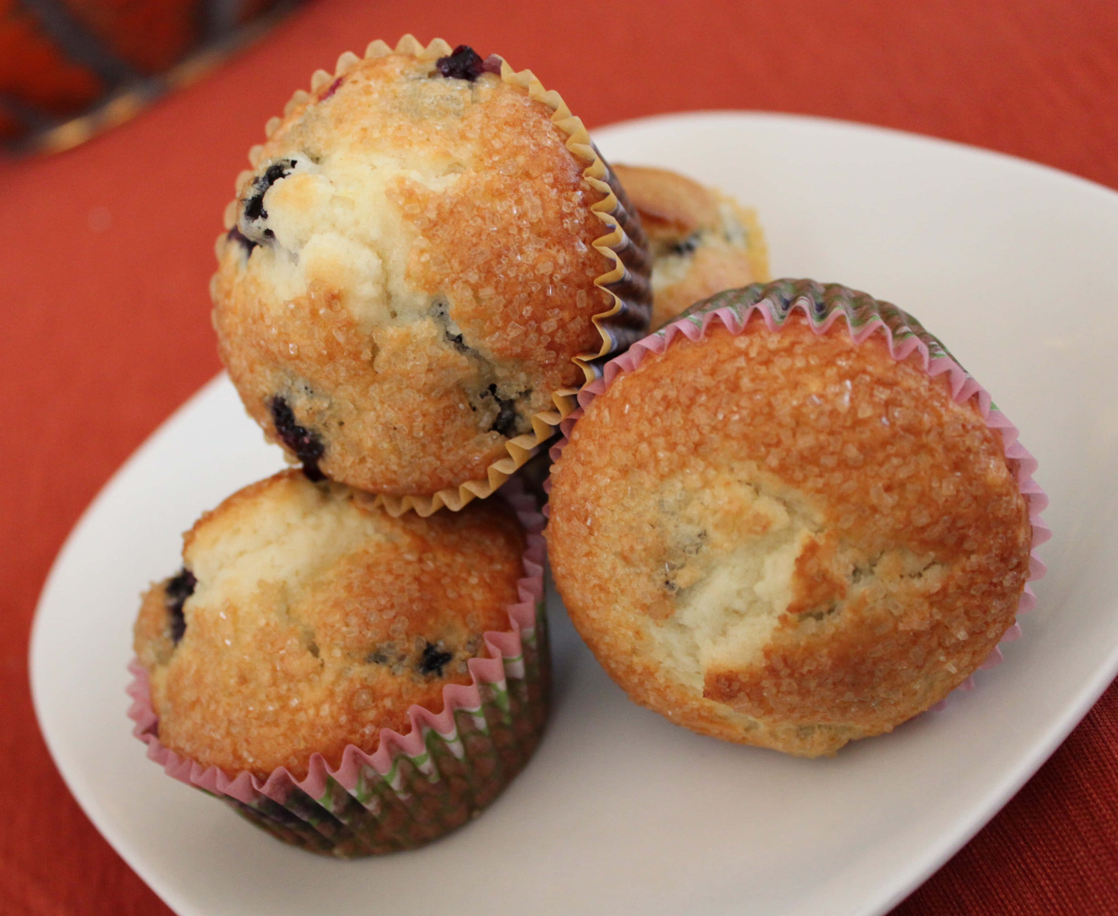 Blueberry Muffins 桌布 壁紙 — 三個金黃色藍莓鬆餅放在彩色紙托中，堆疊在白色碟子上，背景為紅色布料。