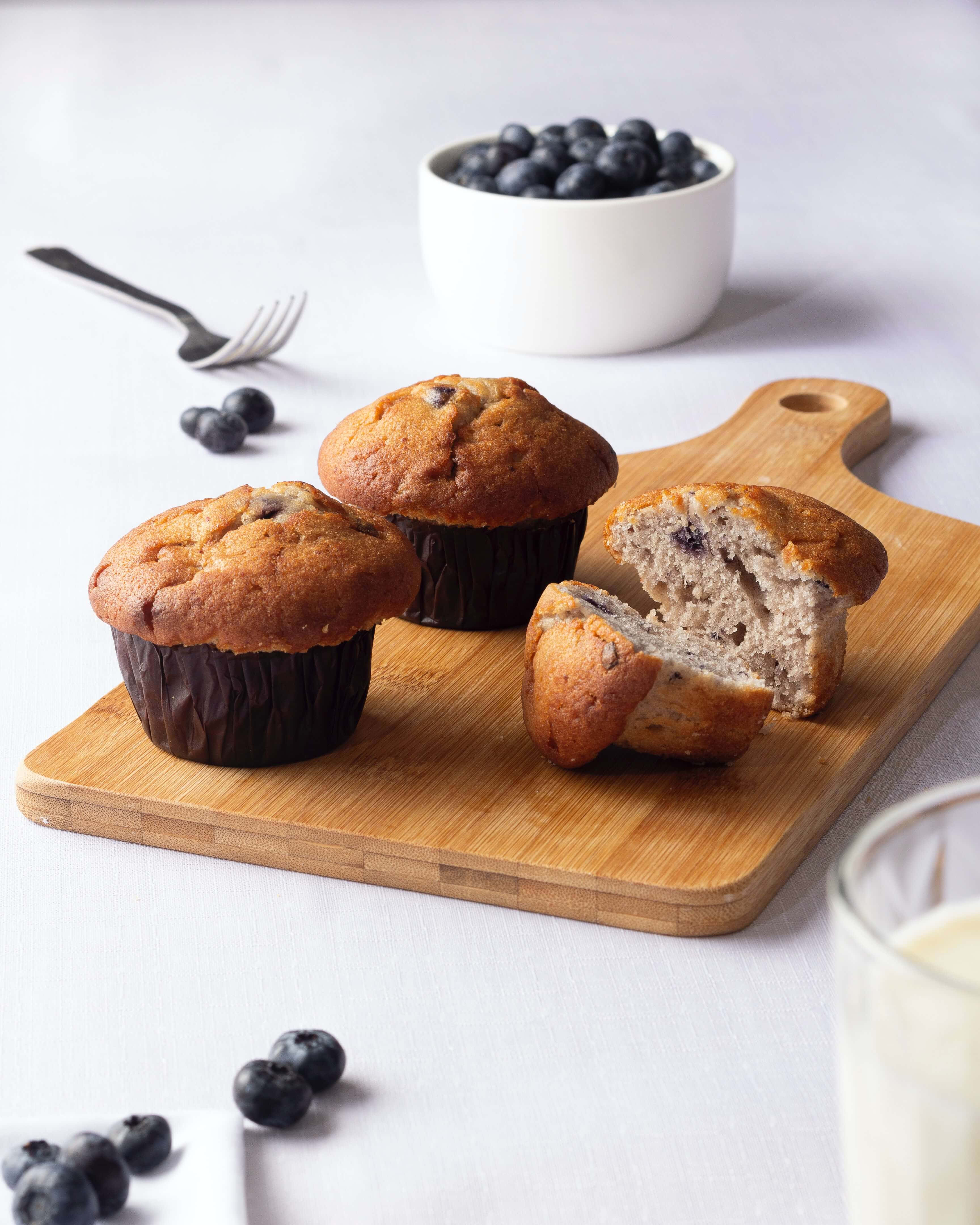 Blueberry Muffins 桌布 壁紙 — 三個金黃色藍莓鬆餅放在木板上，其中一個切開露出濕潤的碎屑，散落著新鮮藍莓，旁邊有一杯牛奶。