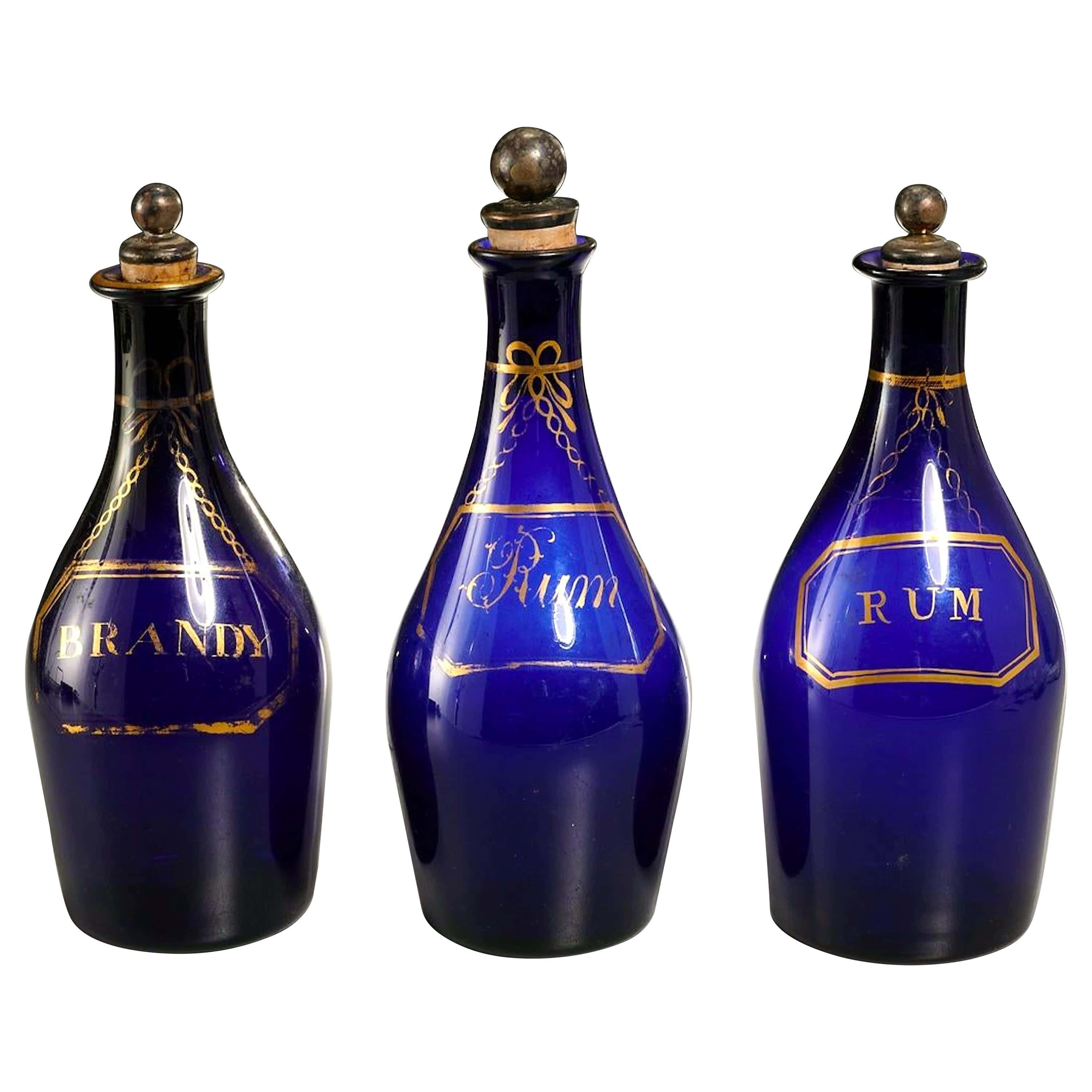 Cobalt Blue 桌布 壁紙 — 三個鈷藍色玻璃醒酒瓶，帶有金色字母和瓶蓋，標有白蘭地朗姆酒和朗姆酒，立在白色背景前。