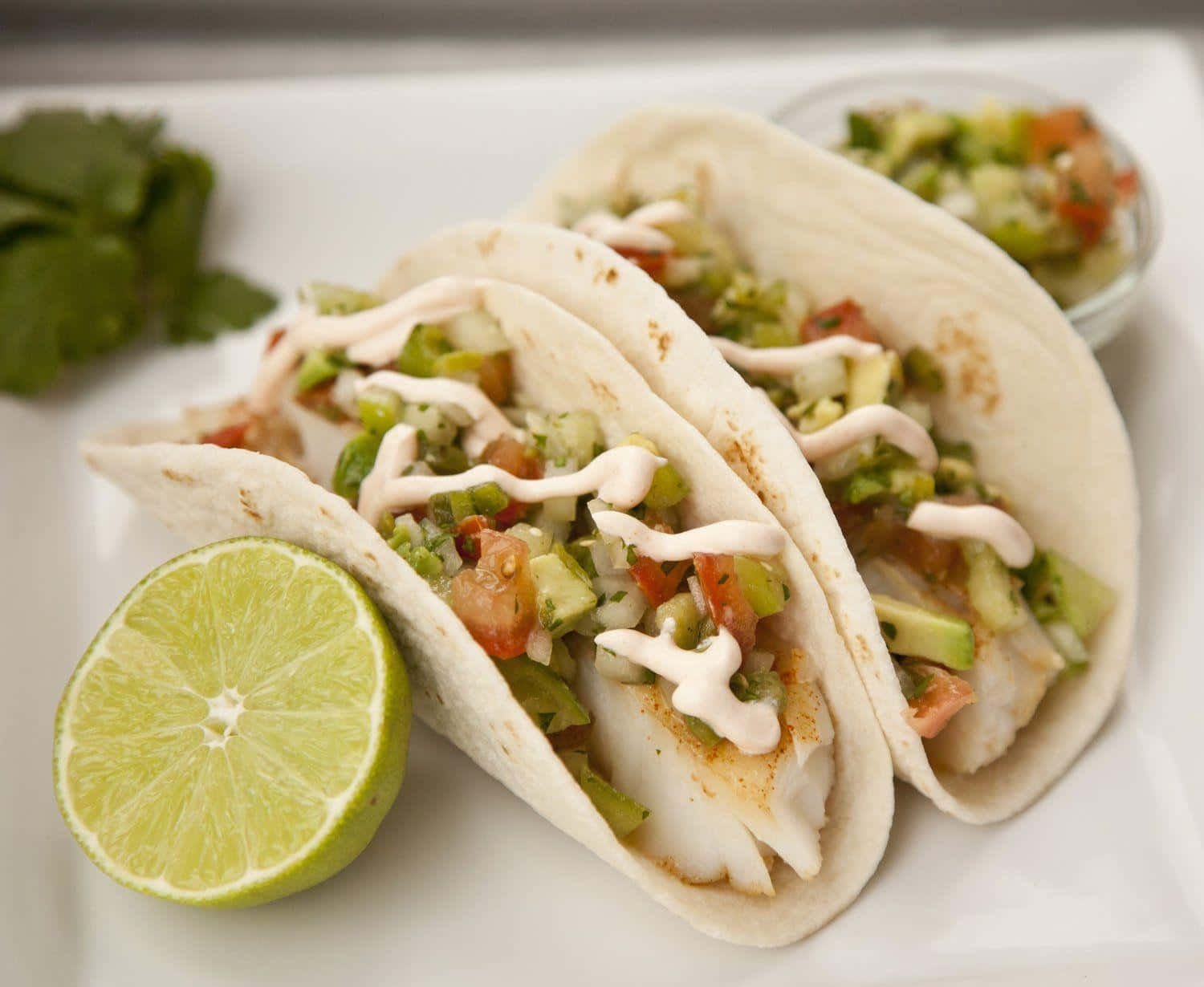 Taco 桌布 壁紙 — 三個魚肉玉米餅配新鮮莎莎醬和奶油醬汁，旁邊放著青檸角和芫荽，置於白色碟上。