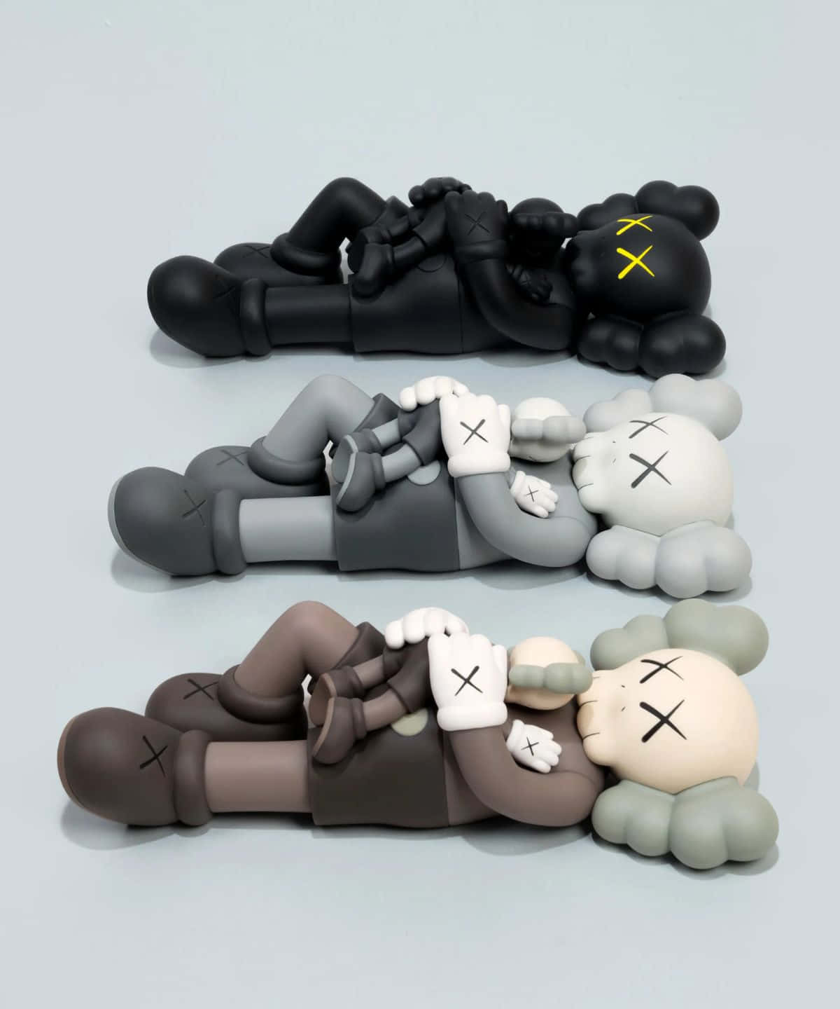 Kaws Figures 桌布 壁紙 — 三個黑色、灰色和棕色的KAWS Companion公仔，帶有X形眼睛和雲朵頭部，堆疊在純淺灰色表面上。