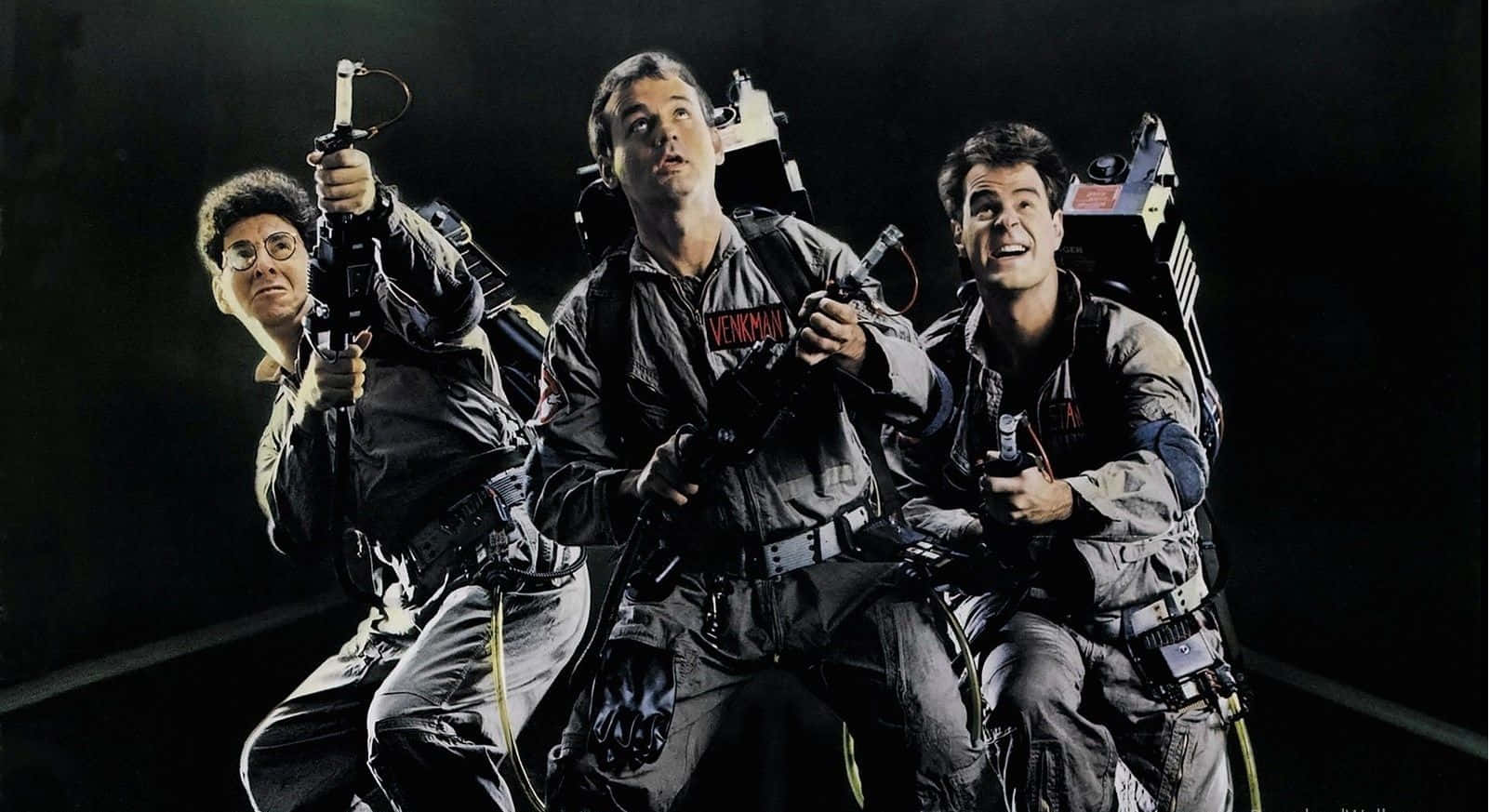 Ghostbusters 桌布 壁紙 — 三名捉鬼敢死隊穿着標誌性裝備，在黑暗背景中帶着發光的質子背包動態擺姿勢。