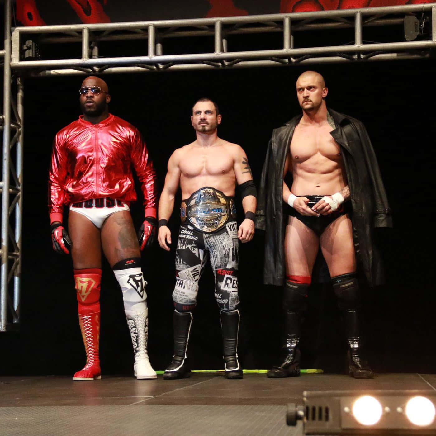 三名摔角手 Austin Aries、Killer Kross 與 Moose 在 Impact Wrestling 2018 的精彩合影 壁紙 — 三位肌肉發達的摔角手身穿大膽服裝，在明亮燈光下自信地站在舞台上，手持冠軍腰帶。