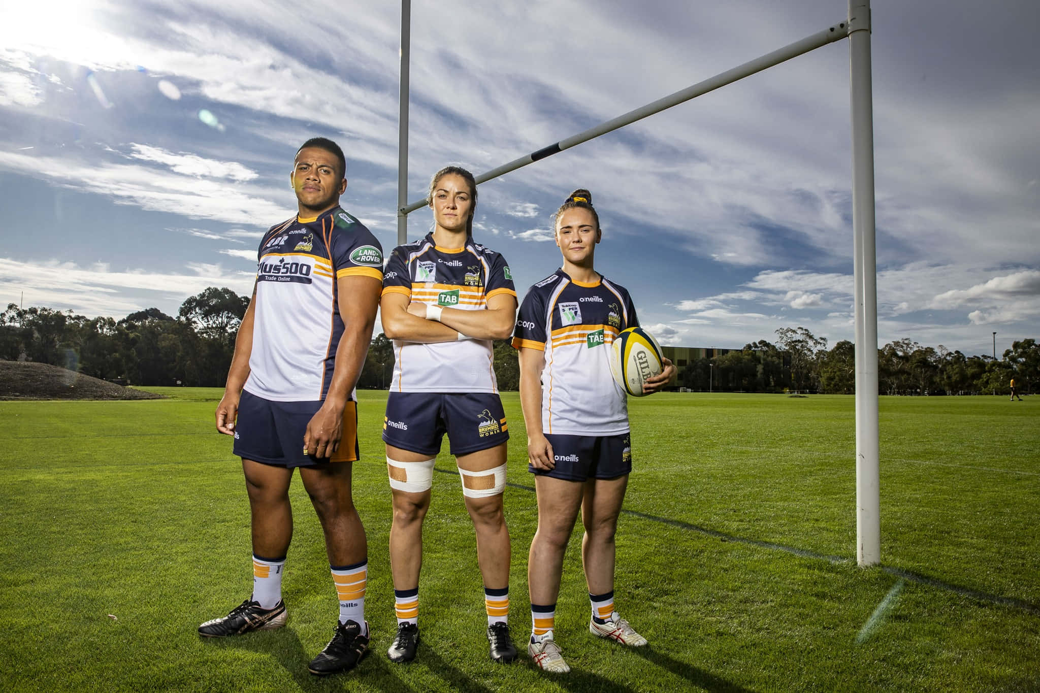 Brumbies 桌布 壁紙 — 三名欖球運動員身穿隊服，自信地站在草地上，身後是部分多雲的天空和球門柱。