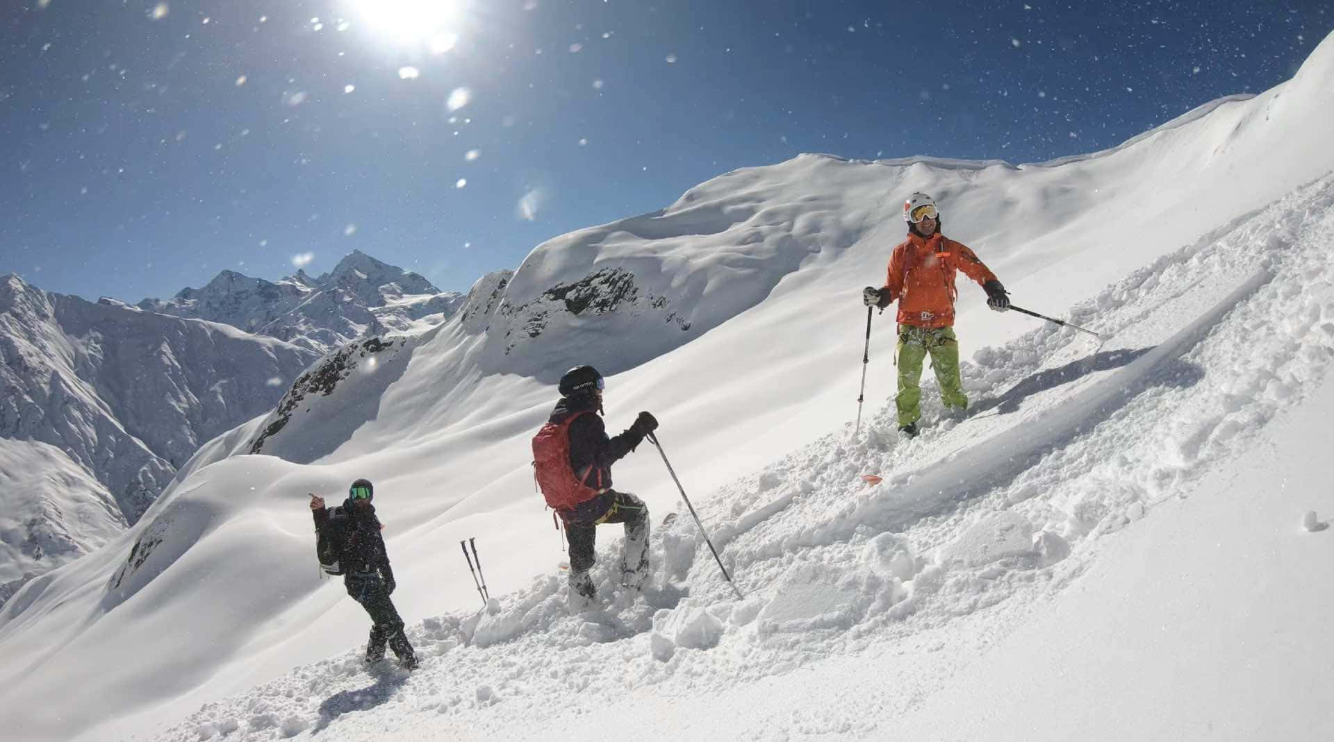 Skiing 桌布 壁紙 — 三名滑雪者在晴朗藍天和太陽耀斑下，於明亮的雪山斜坡上滑行。
