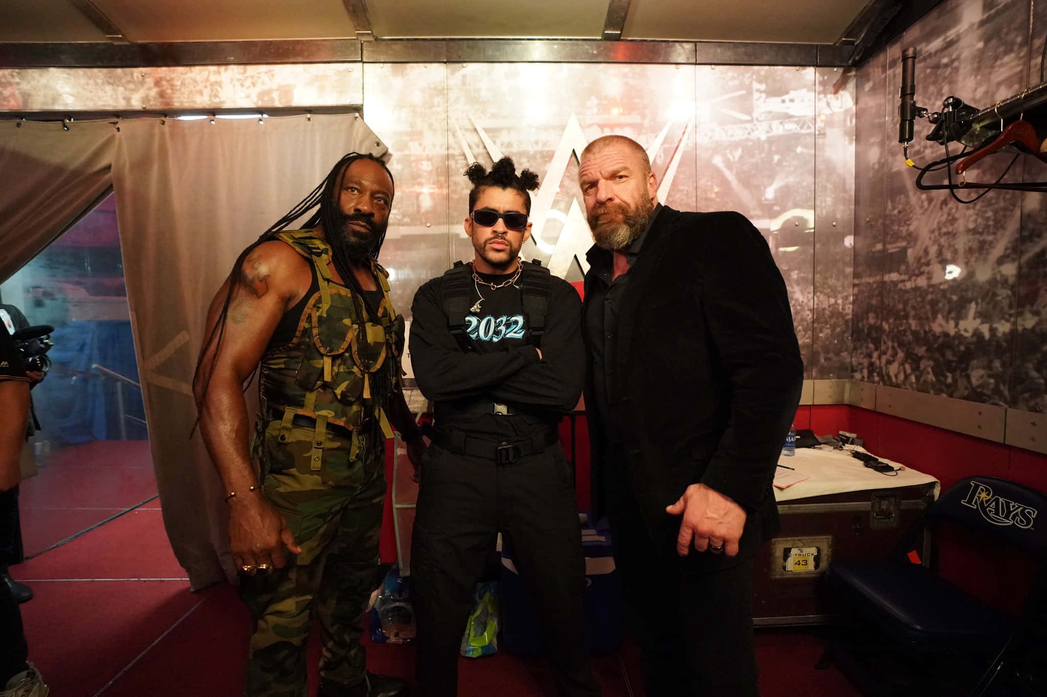 Booker T With Bad Bunny And Triple H 壁紙 — 三名男子在後台摔角區域自信地擺姿勢，該區域有紅色地板和一個「RAYS」行李箱在附近。