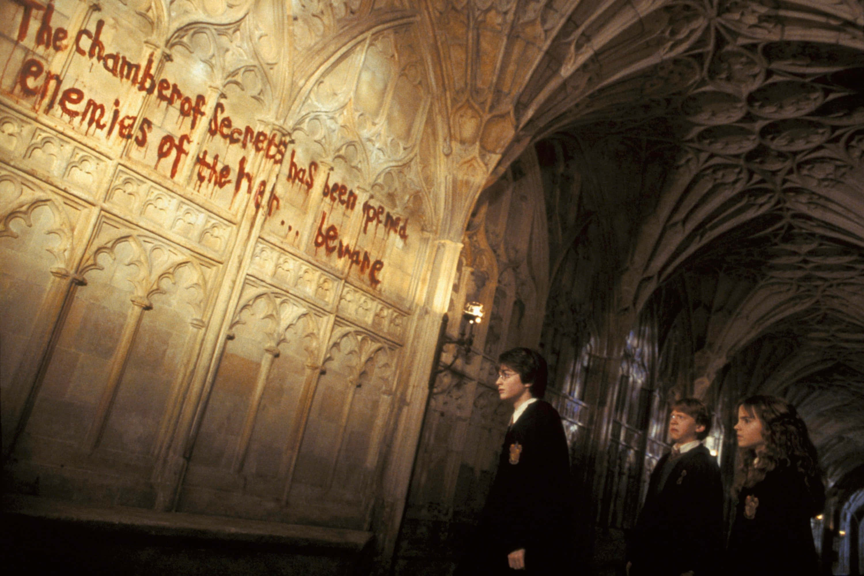The Chamber Of Secrets 桌布 壁紙 — 三名穿著霍格華茲長袍的學生在華麗的石拱門下敬畏地站著，發光的血紅色文字警告著密室。