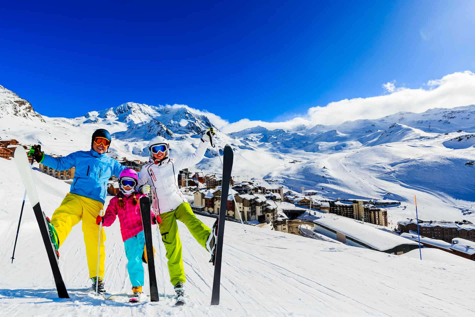 Skiing 桌布 壁紙 — 三名穿著鮮豔裝備的滑雪者，在晴朗藍天下，於白雪覆蓋的山坡頂端愉快地擺姿勢，下方是灑滿白雪的村莊。