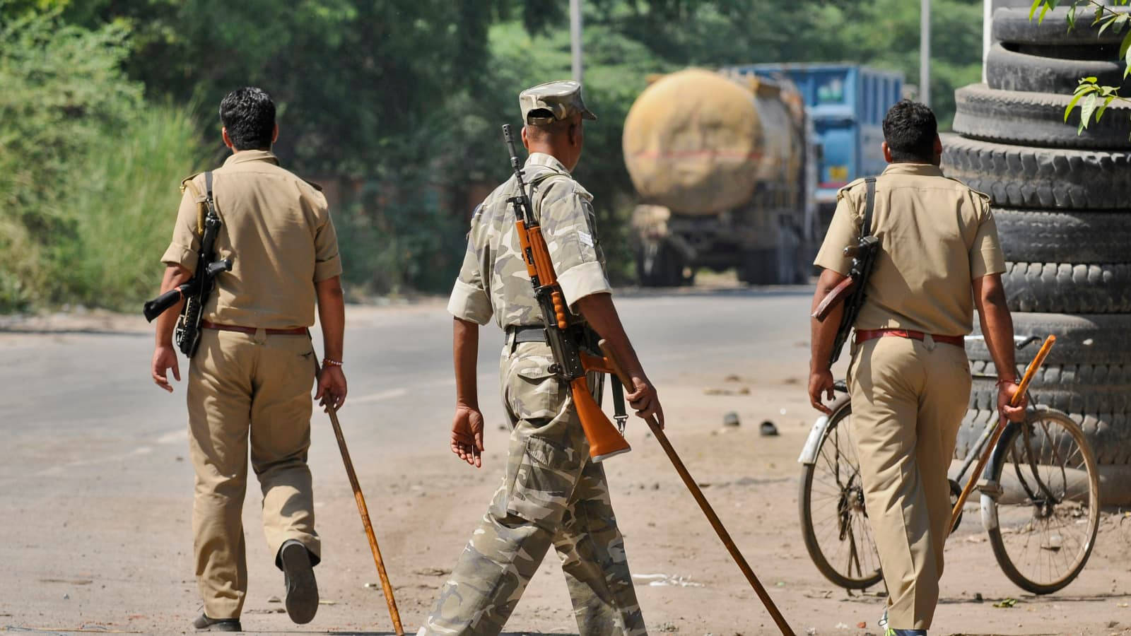 Indian Army 桌布 壁紙 — 三名身穿卡其色制服嘅武裝警察離開鏡頭，沿住一條塵土飛揚嘅路邊行，背景有油罐車同堆疊嘅輪胎。
