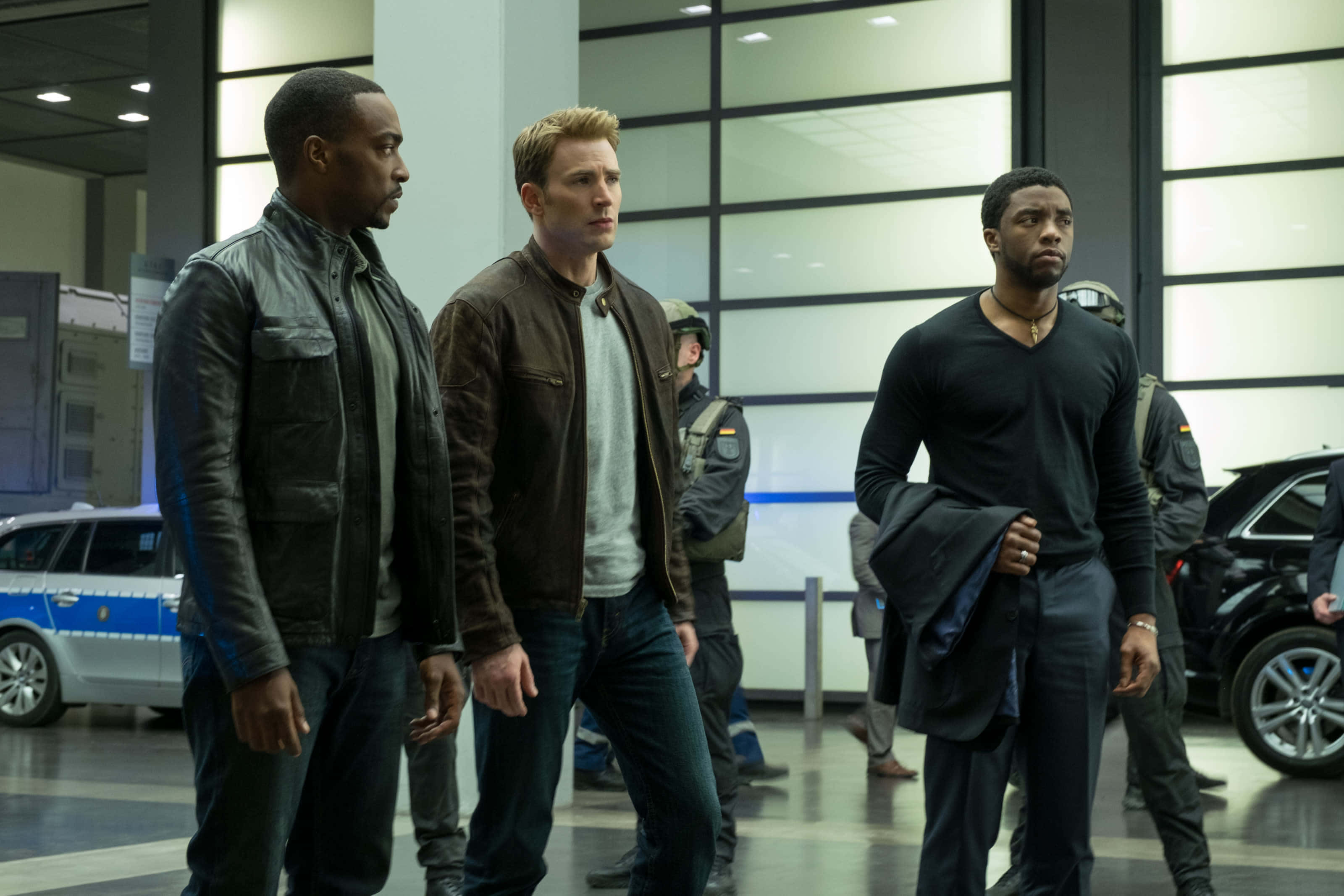 Mcu Characters 桌布 壁紙 — 三名身穿外套的男子在現代機場航站樓內保持警覺，附近有警車和保安人員。