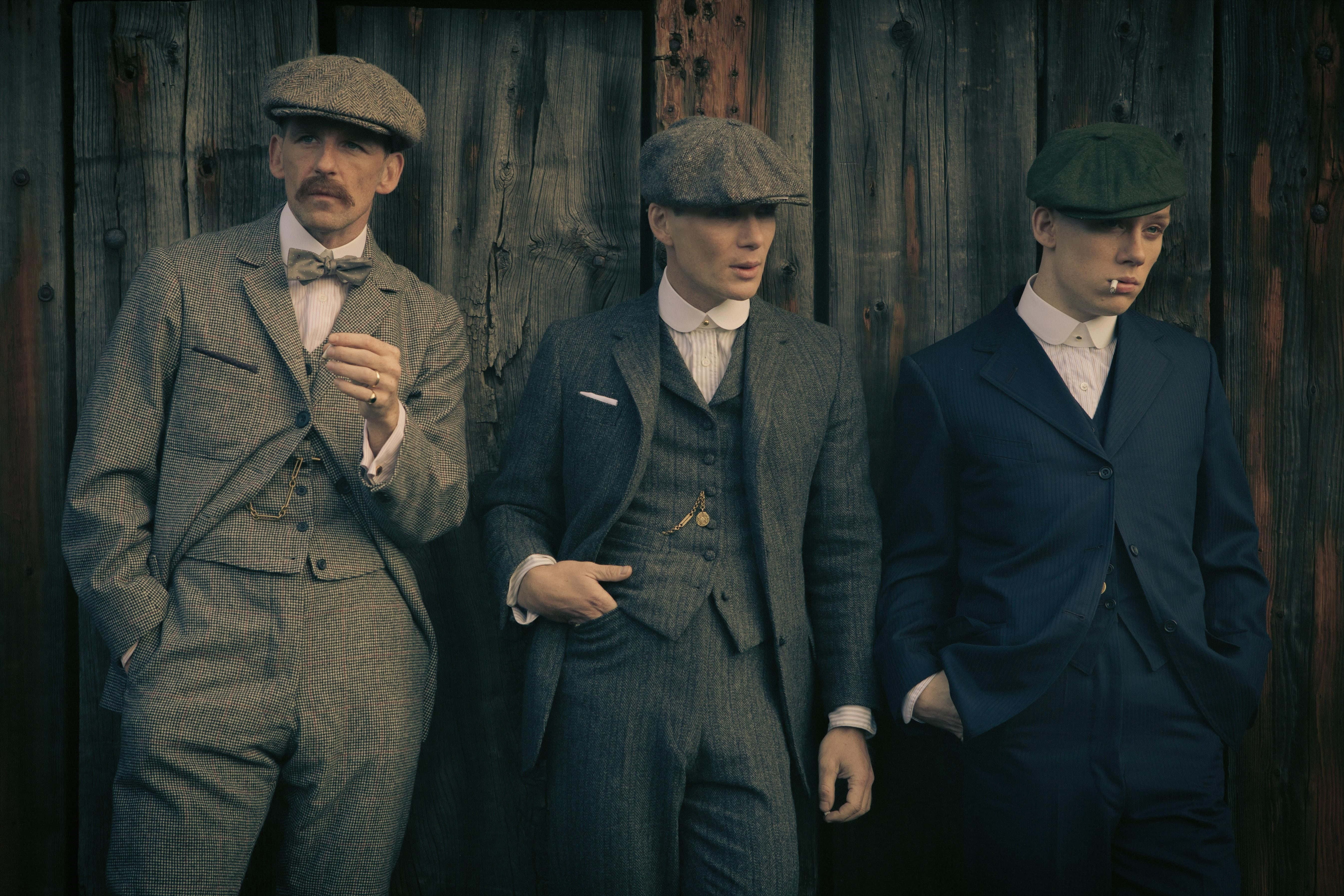 Peaky Blinders 桌布 壁紙 — 三名身穿復古西裝和扁帽的男子，表情嚴肅地靠在一面風化的木牆上。