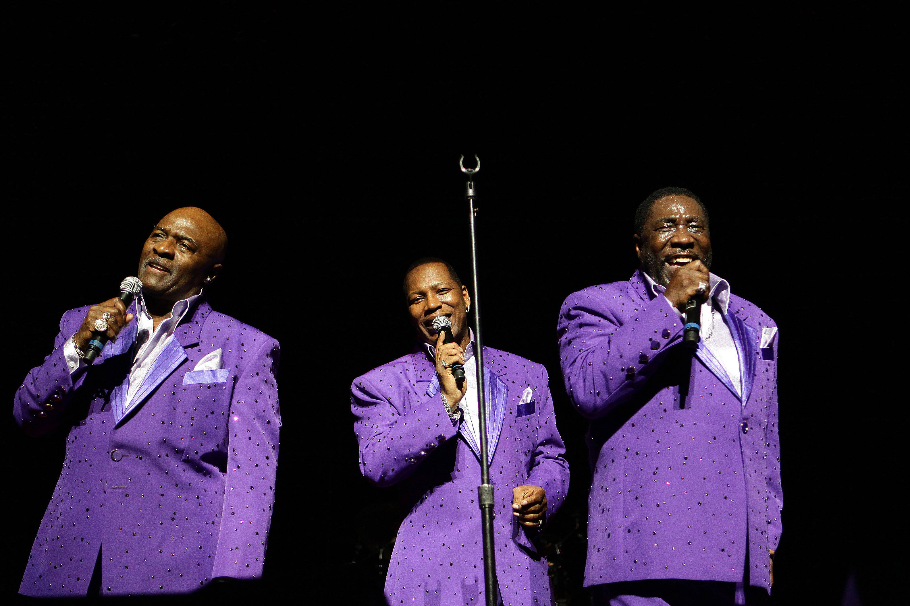 Walter Williams Ojays《We Cried Together》 壁紙 — 三名身穿閃亮紫色西裝的男子在黑色背景的舞台燈光下對著麥克風唱歌。