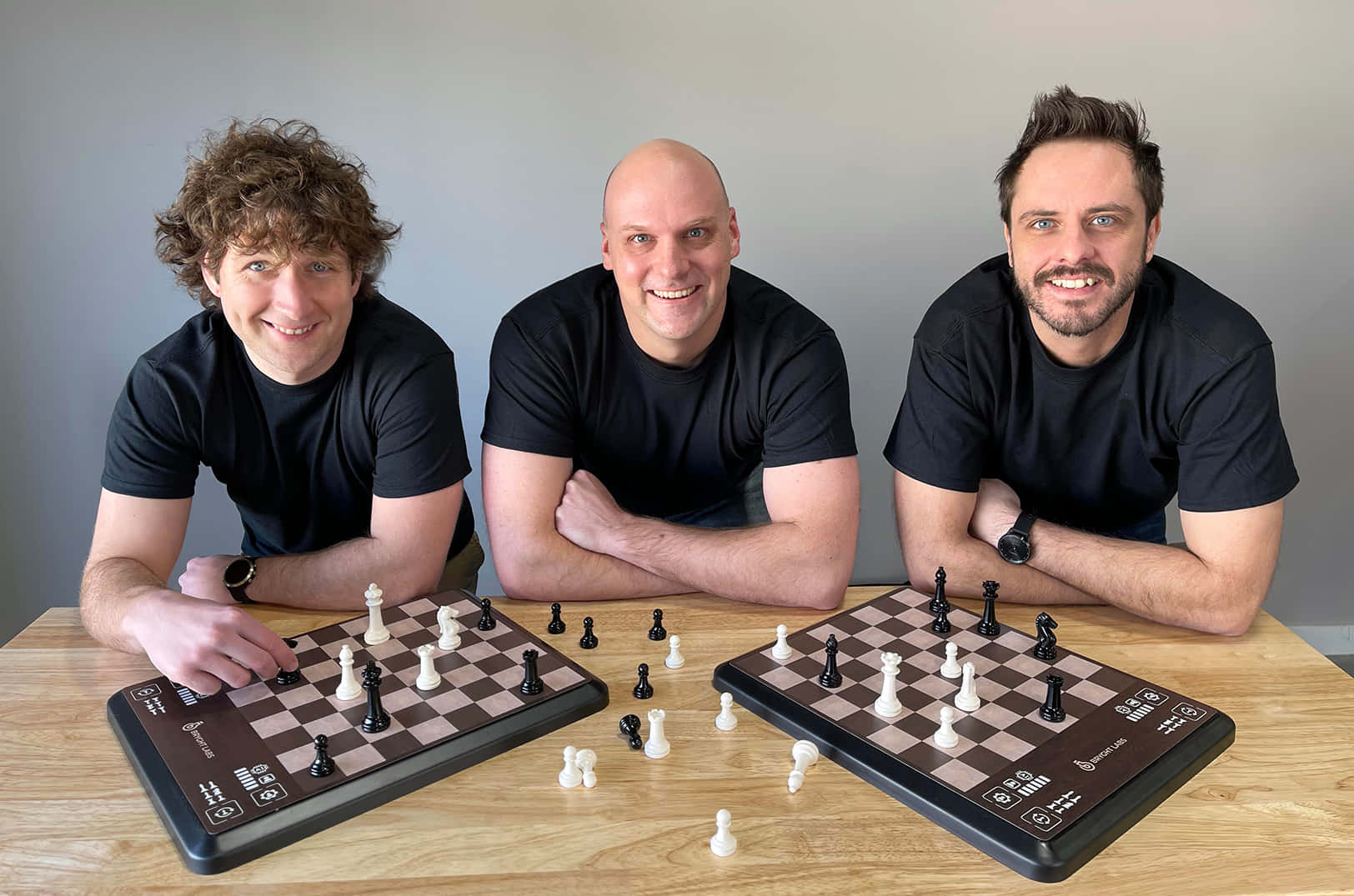 Chessboard 桌布 壁紙 — 三名身穿黑色襯衫、面帶微笑的男子，靠在一張張木桌上，桌上放著棋盤和散落的棋子，背景是樸素的灰色牆壁。