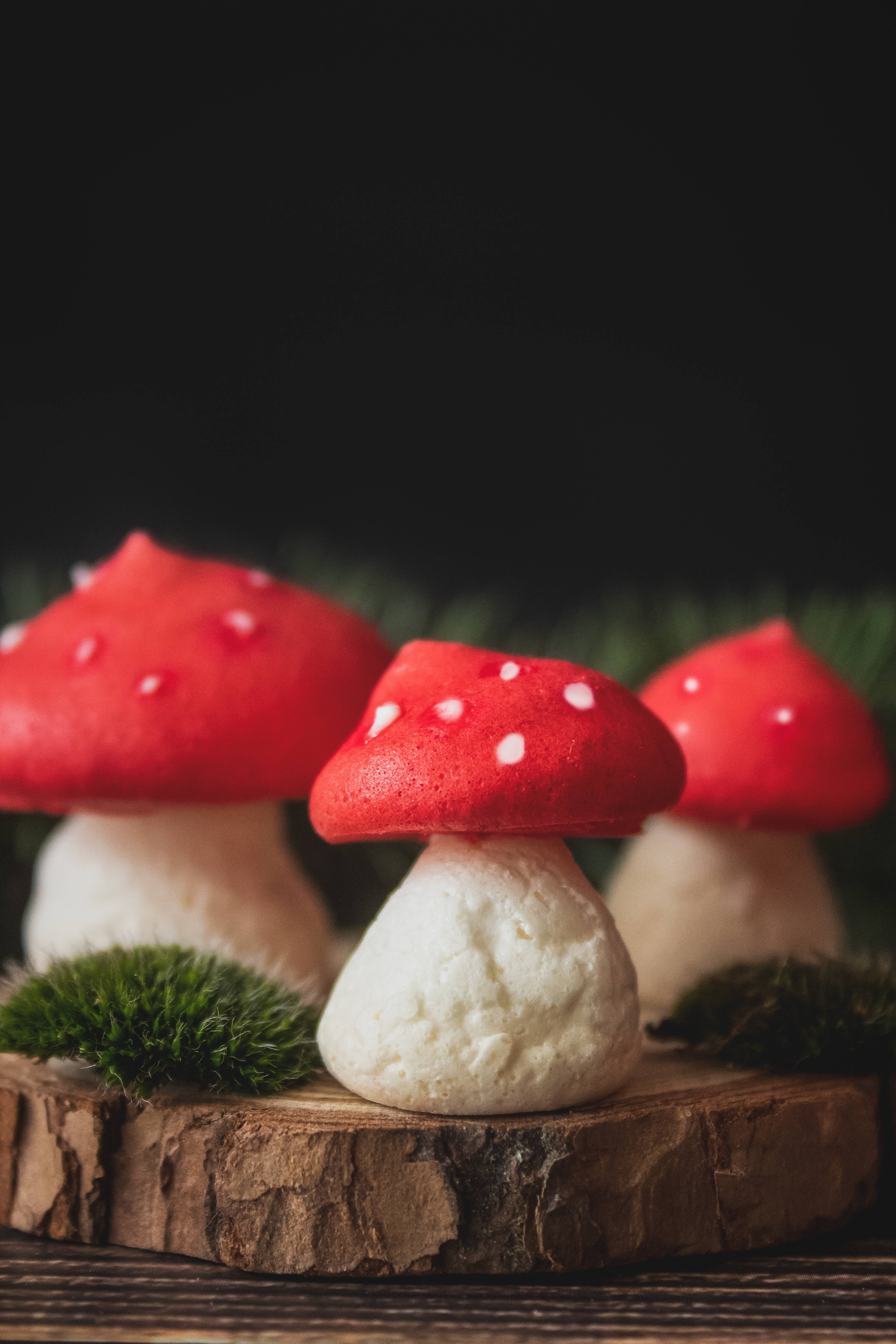 三塊可愛的蘑菇馬林餅 壁紙 — 三個紅白斑點蘑菇甜品放在一塊帶有苔蘚裝飾的鄉村風格木片上，背景深色。