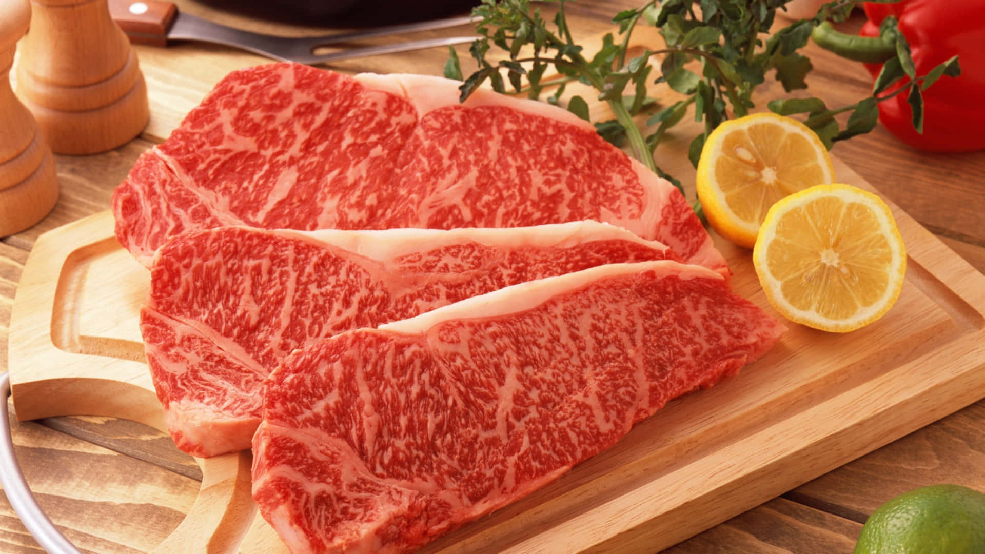 Red Meat 桌布 壁紙 — 三塊大理石紋牛肉扒放在木砧板上，旁邊有檸檬片和新鮮香草。